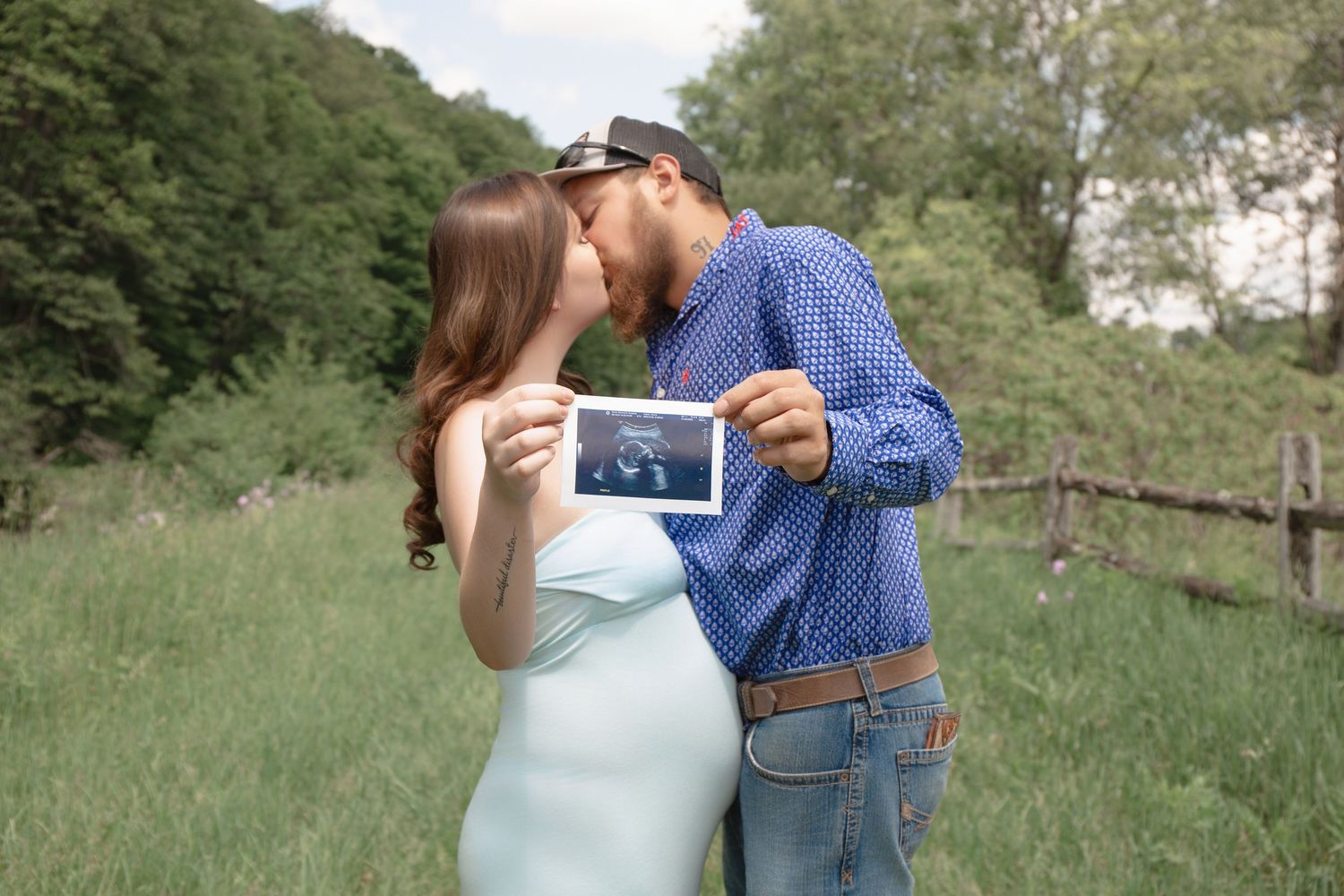 A romantic couple embraces in a sunlit meadow with tall grass and trees.