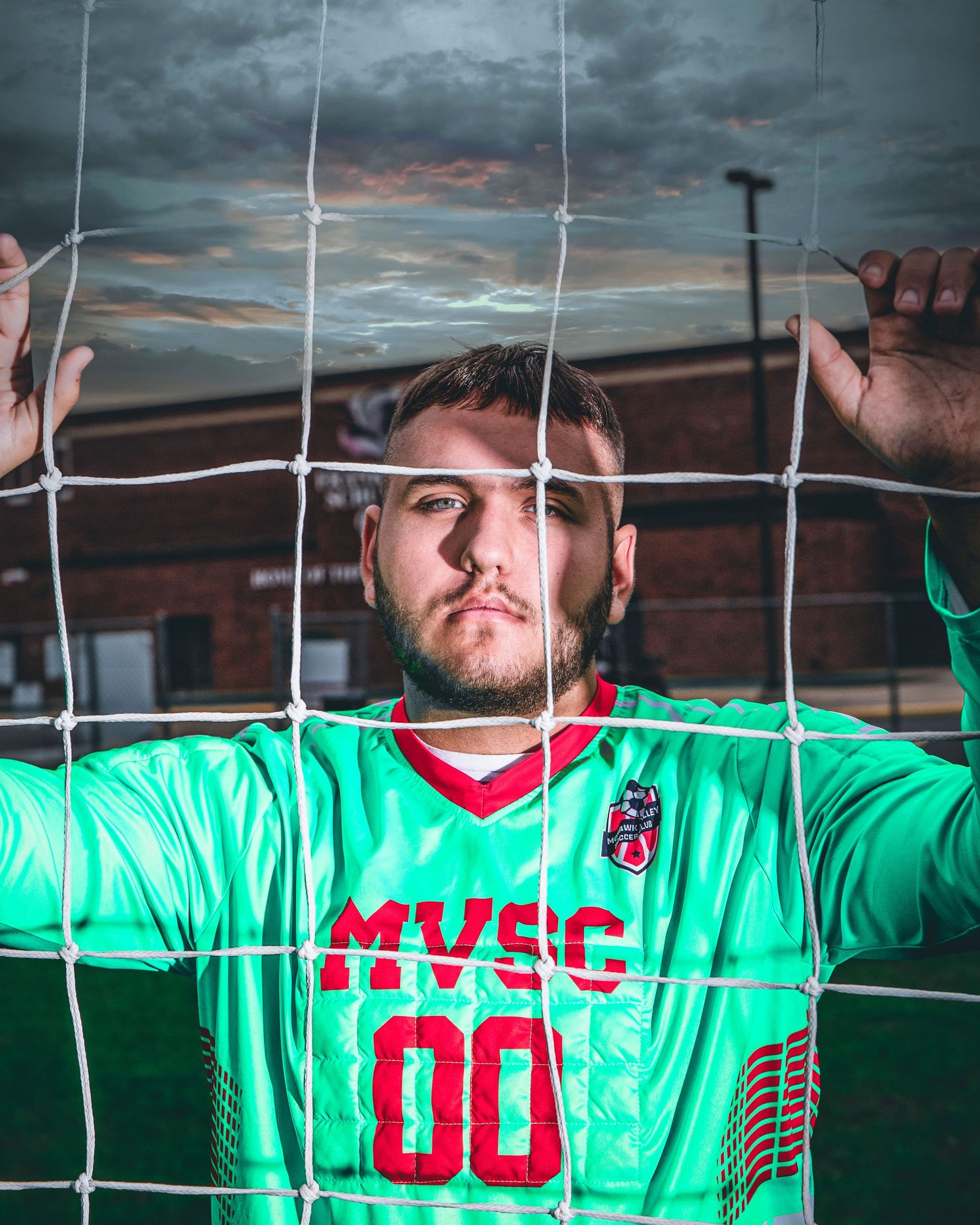 Soccer goalkeeper in green jersey posing against goal netting at sunset.