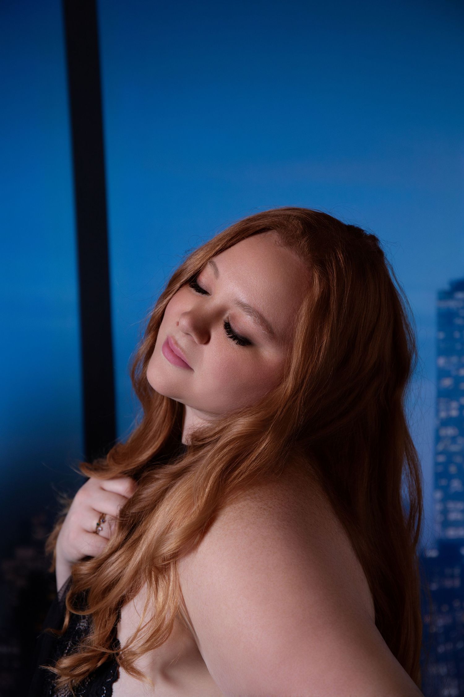 Person with red hair poses against blue evening sky with city buildings visible in background.
