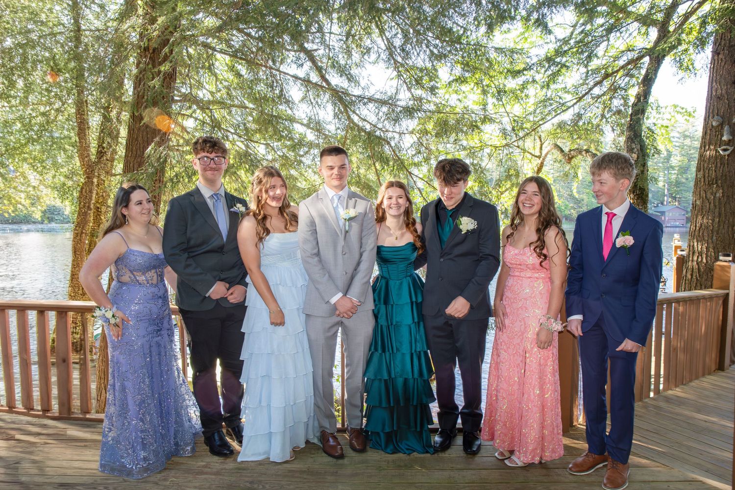 Group of teens in formal prom attire posing on a wooden deck near a lake surrounded by trees.