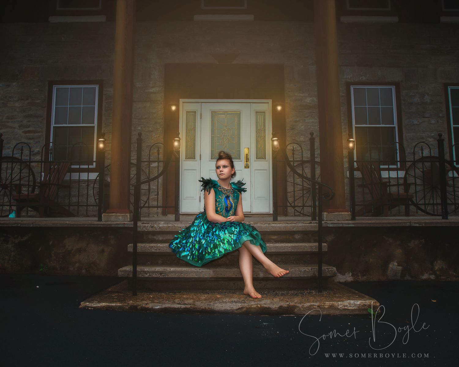 Young girl in teal feathered dress sits on steps of illuminated building at dusk.
