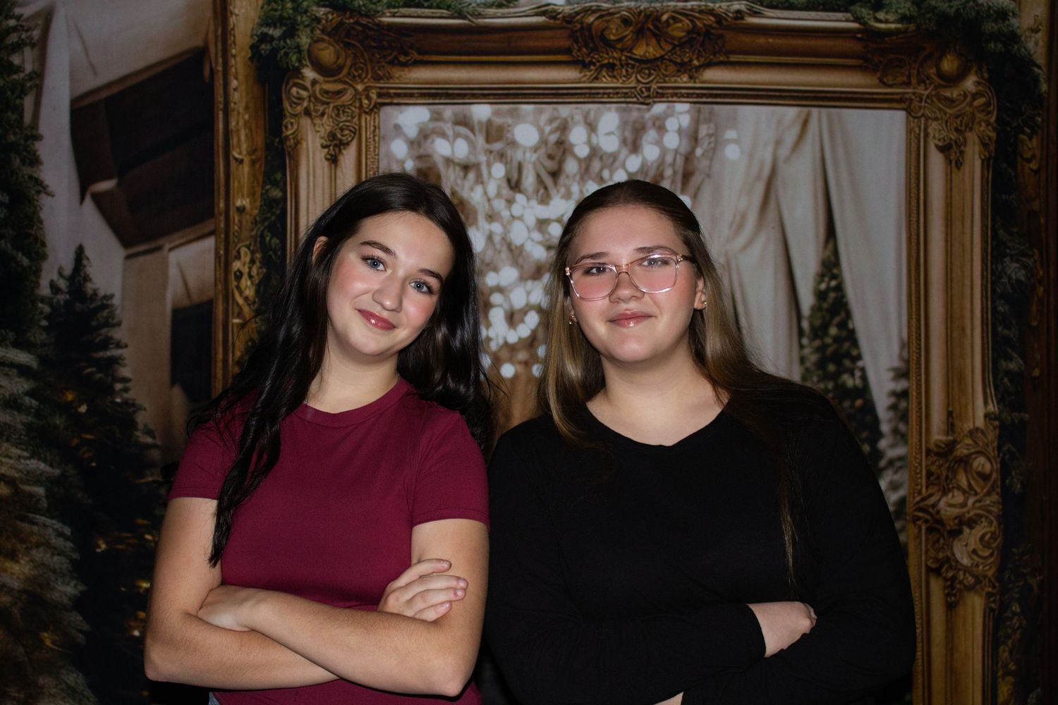 Two people wearing burgundy and black tops pose together in front of an ornate gold mirror and holiday decor.