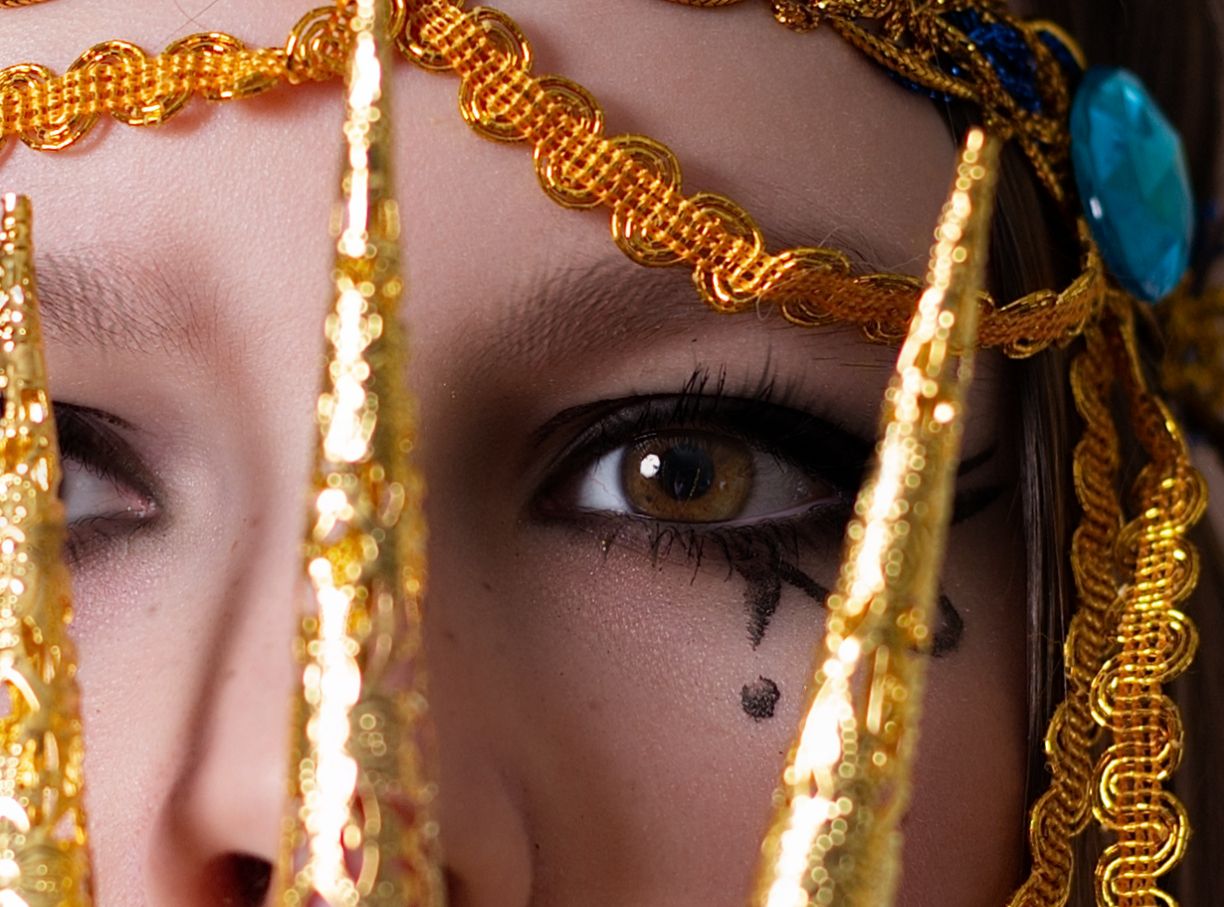 Closeup view through golden tinsel strands showing an eye with dramatic makeup and false eyelashes.