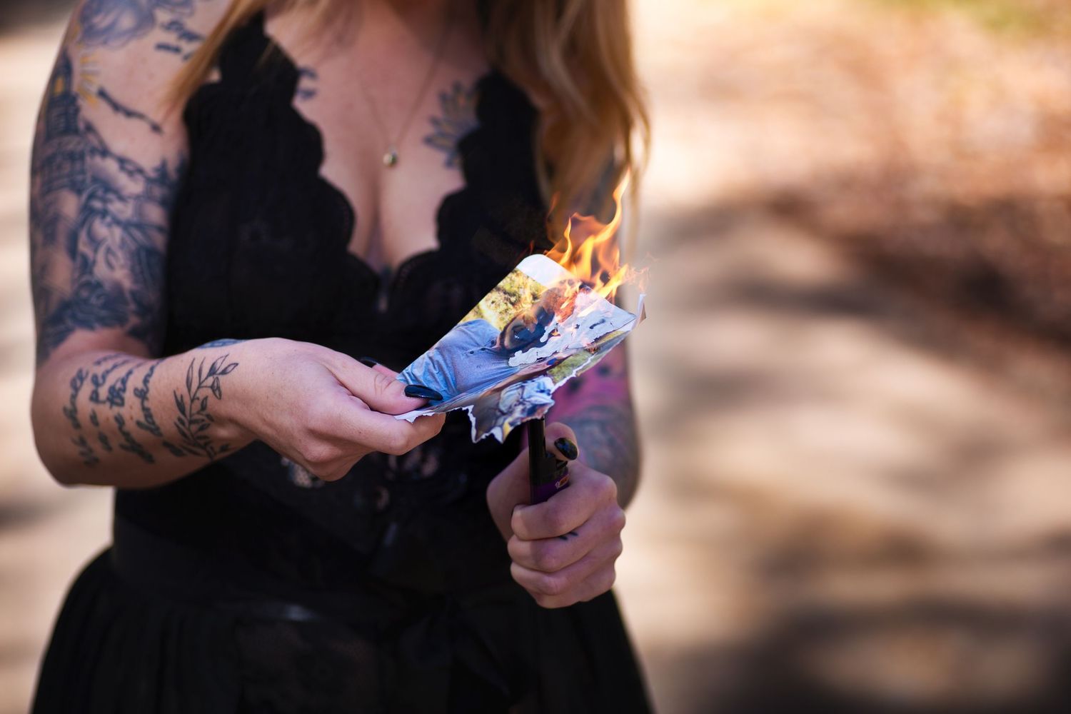 A tattooed hand holds a burning piece of dark purple paper with flames illuminating the edges.