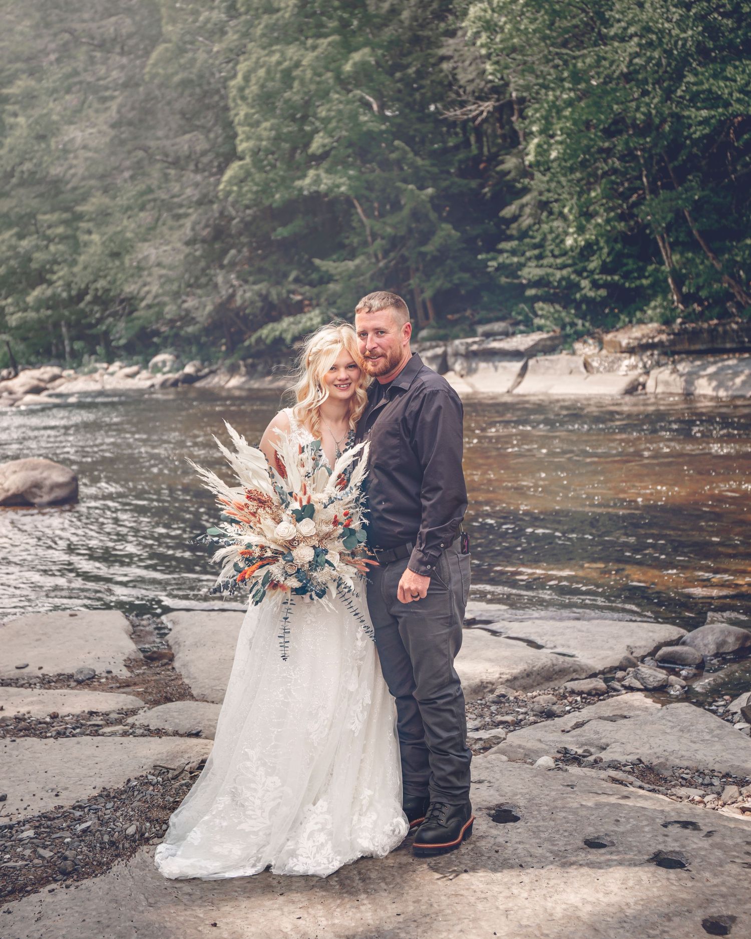 Wedding couple poses on rocky riverbank in natural outdoor setting.