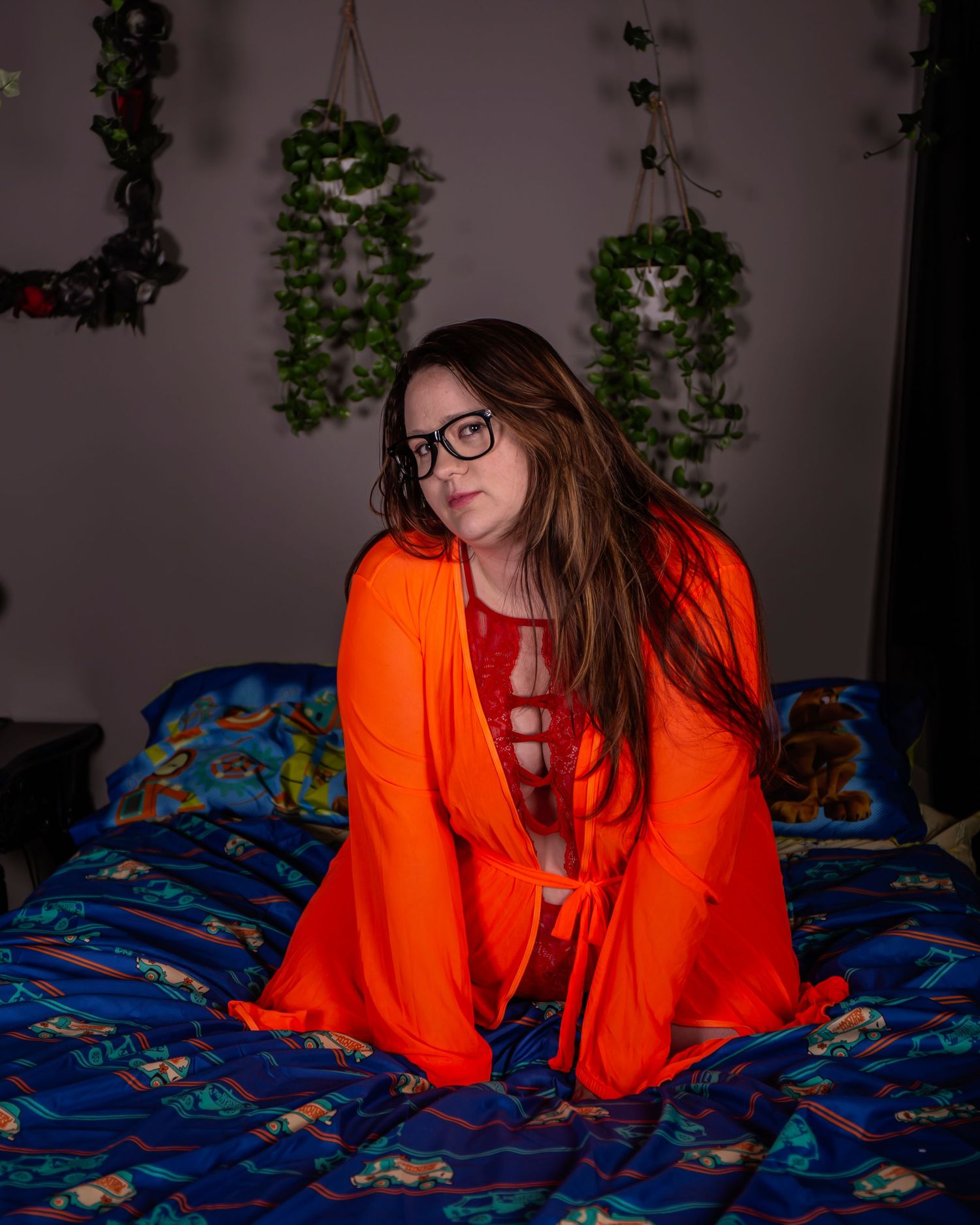 A person in a bright orange sweater reclines on a blue patterned bedspread in a dimly lit room with greenery on the wall.