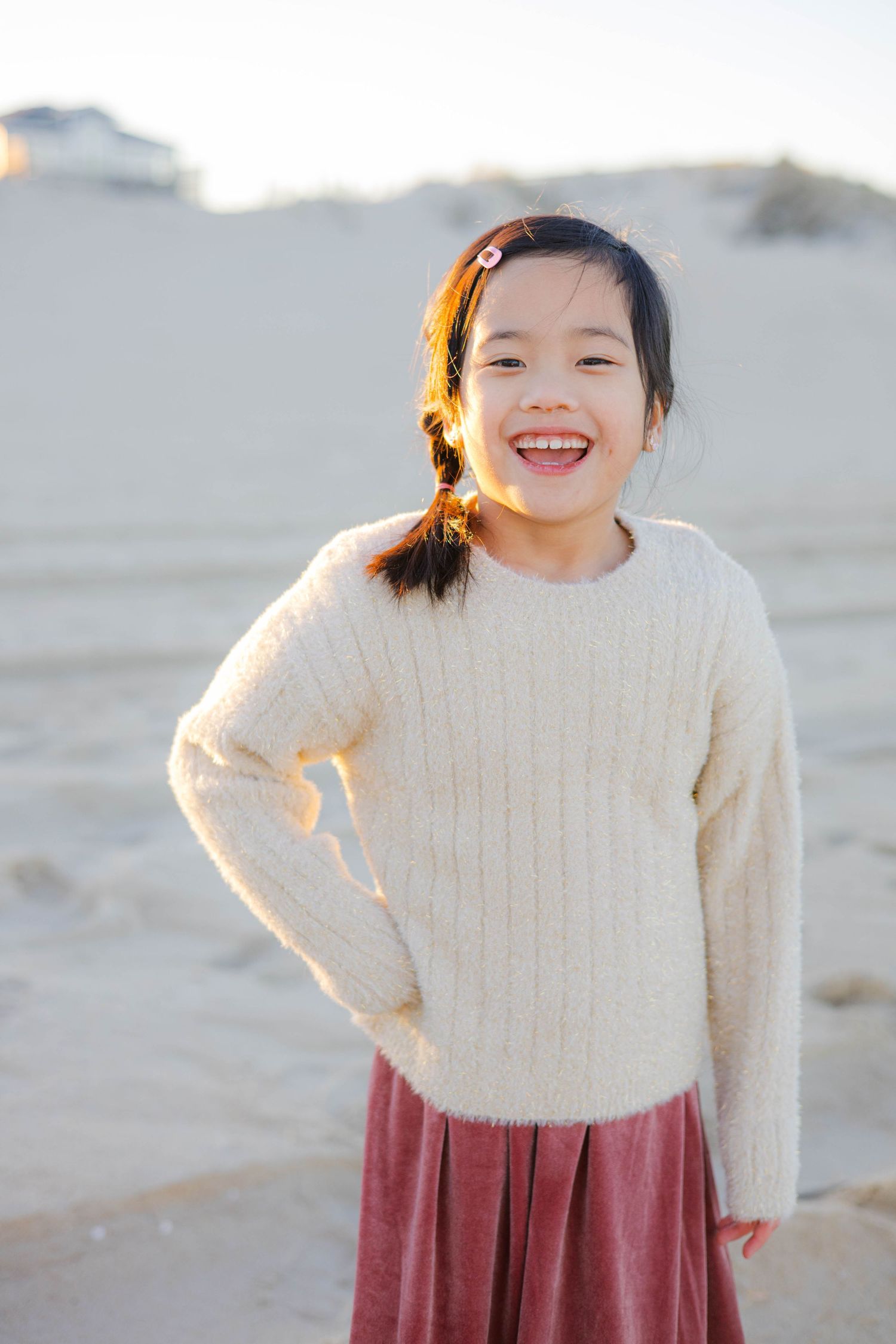 Child in cream sweater and burgundy skirt laughing joyfully on beach.