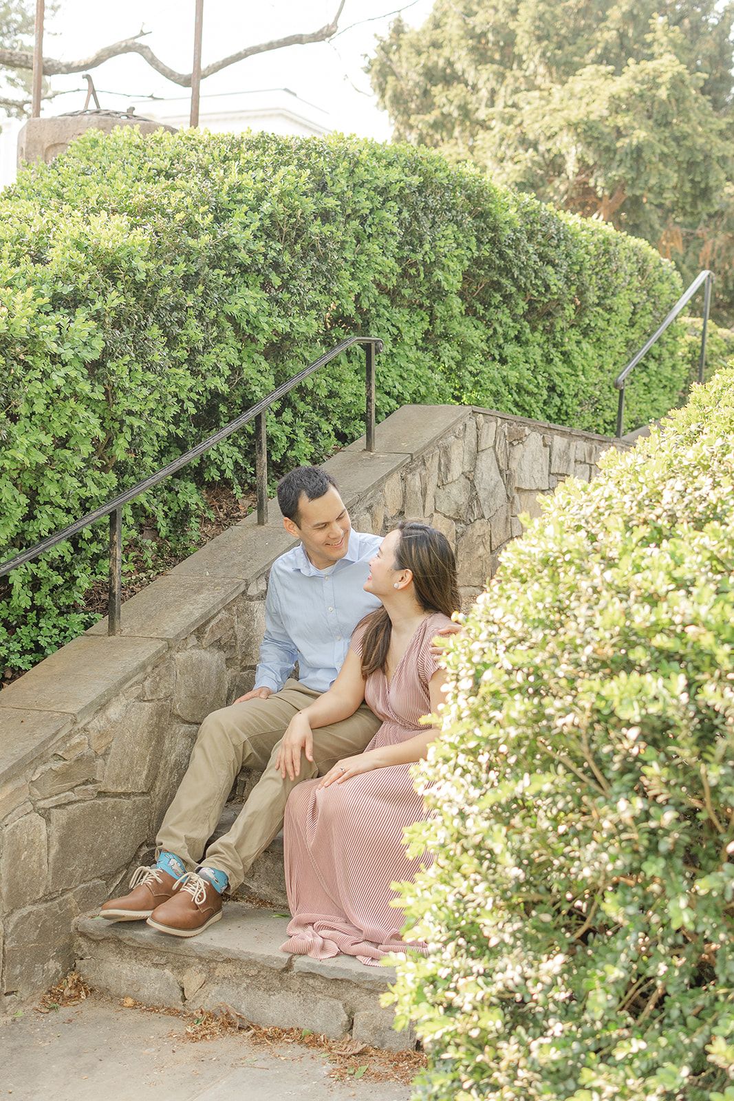 Couple sitting on stone steps surrounded by lush green hedges, woman in pink dress leaning toward man in blue shirt.