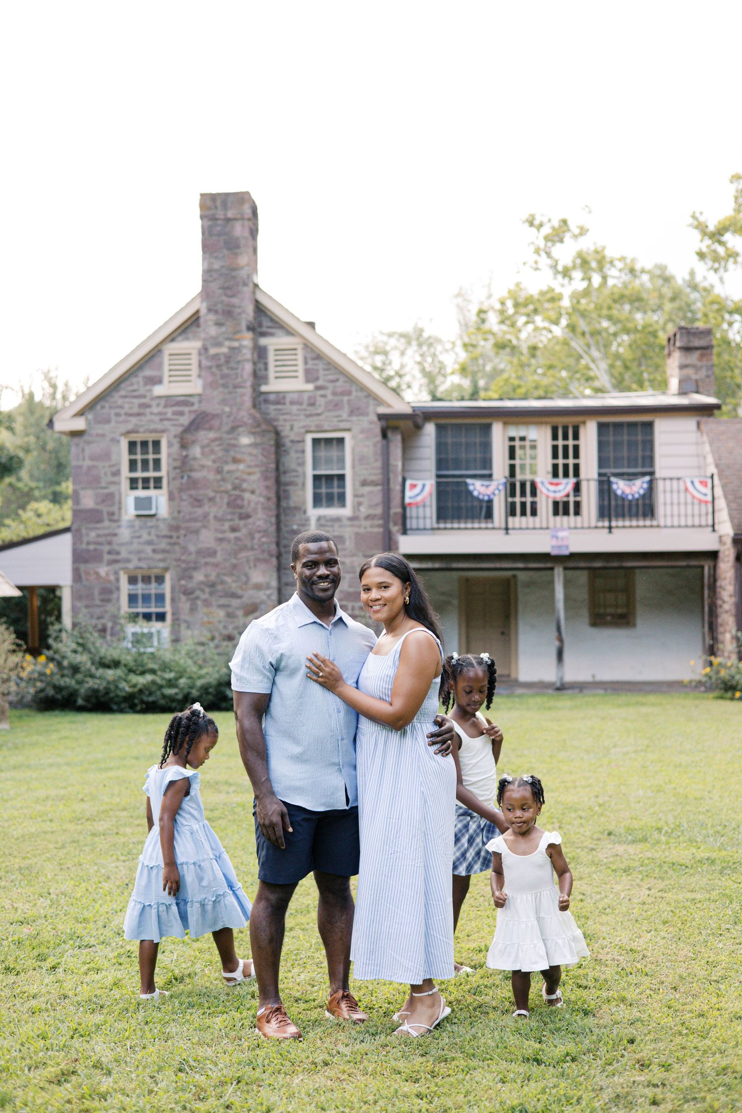 A happy Black family poses together on a green lawn in front of a historic stone house on a sunny day.