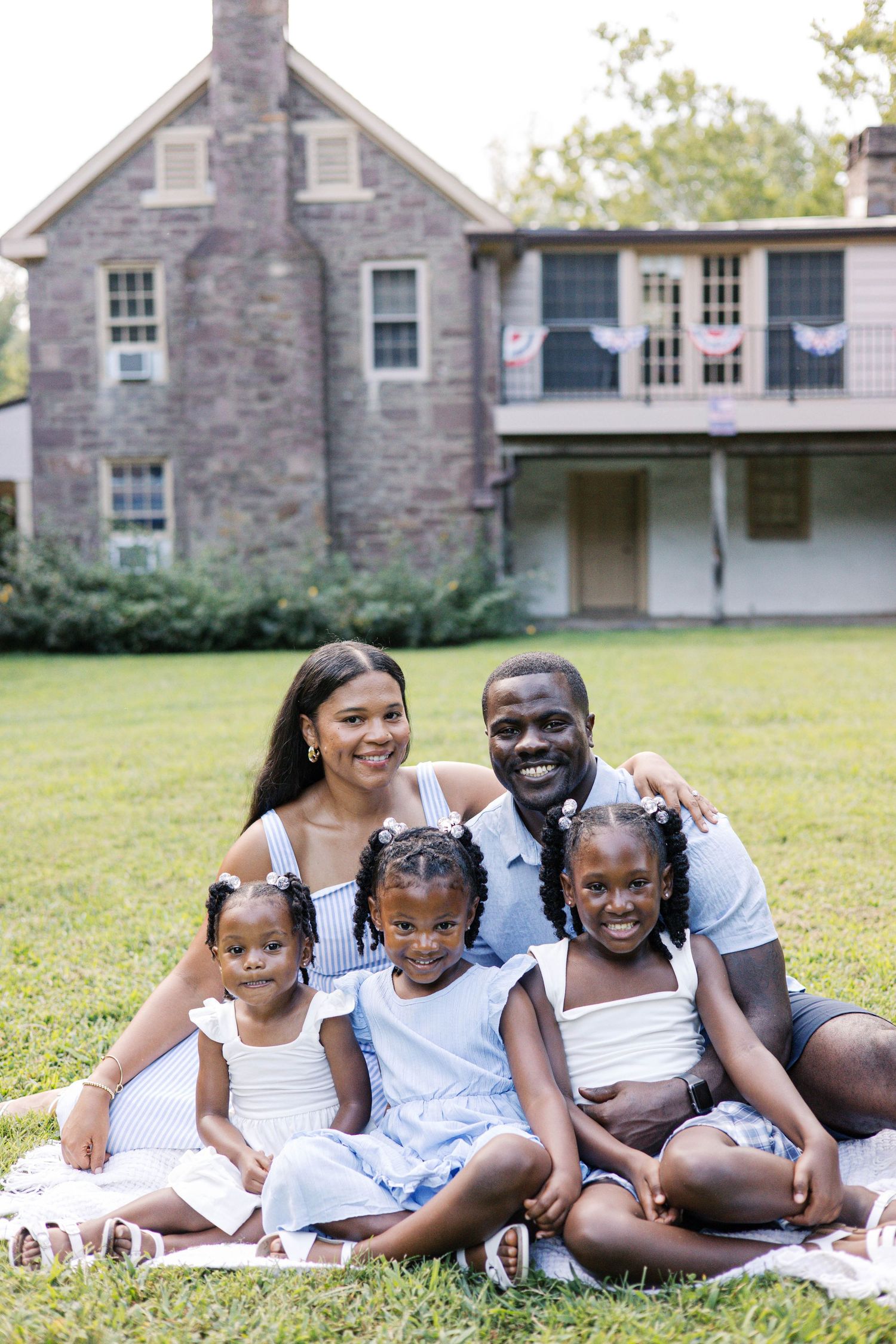 Happy Black family of five posing on a blanket in front of a historic stone house on a green lawn.