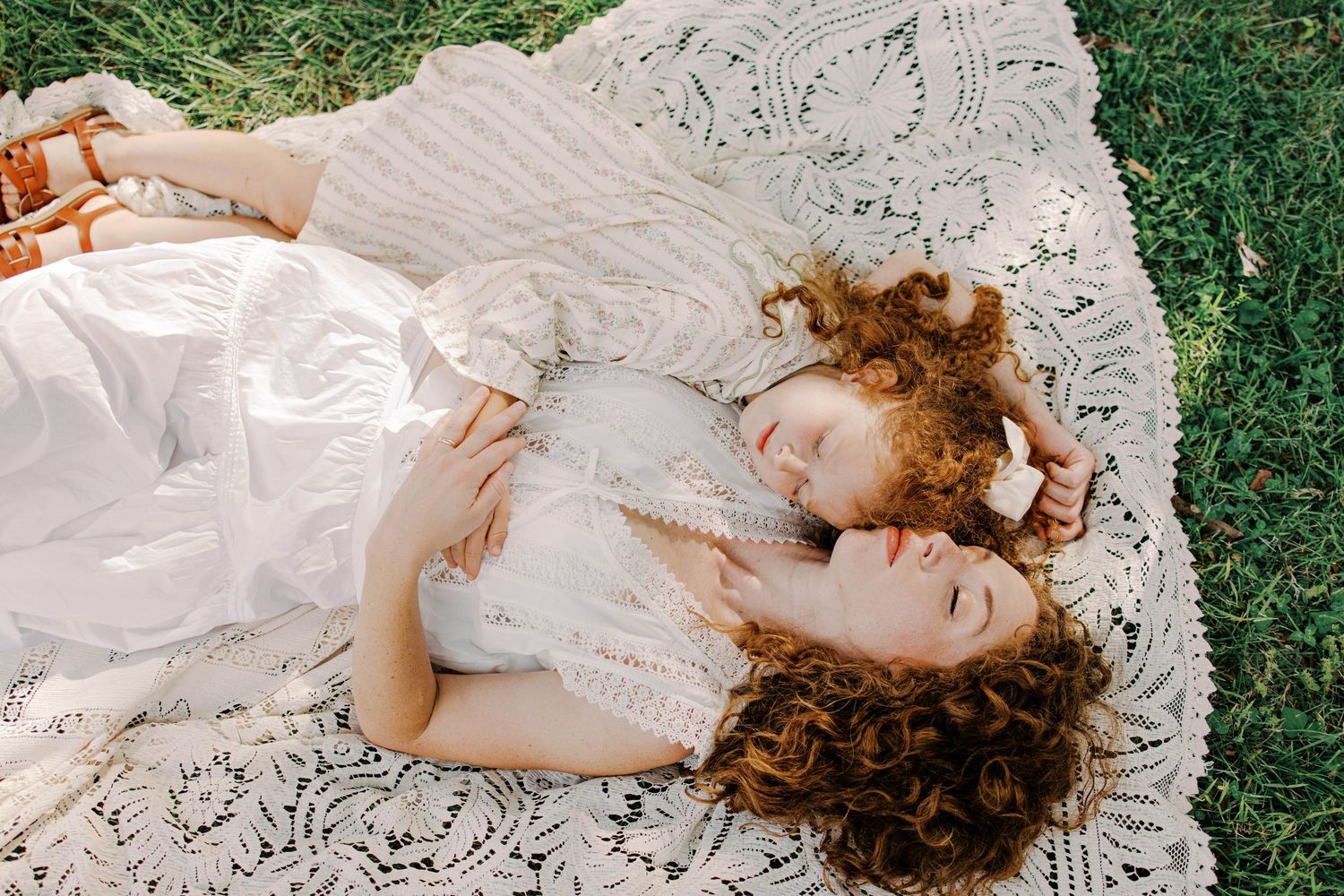 Two curly-haired women in white dresses lie close together on a lace blanket spread across green grass, viewed from above.