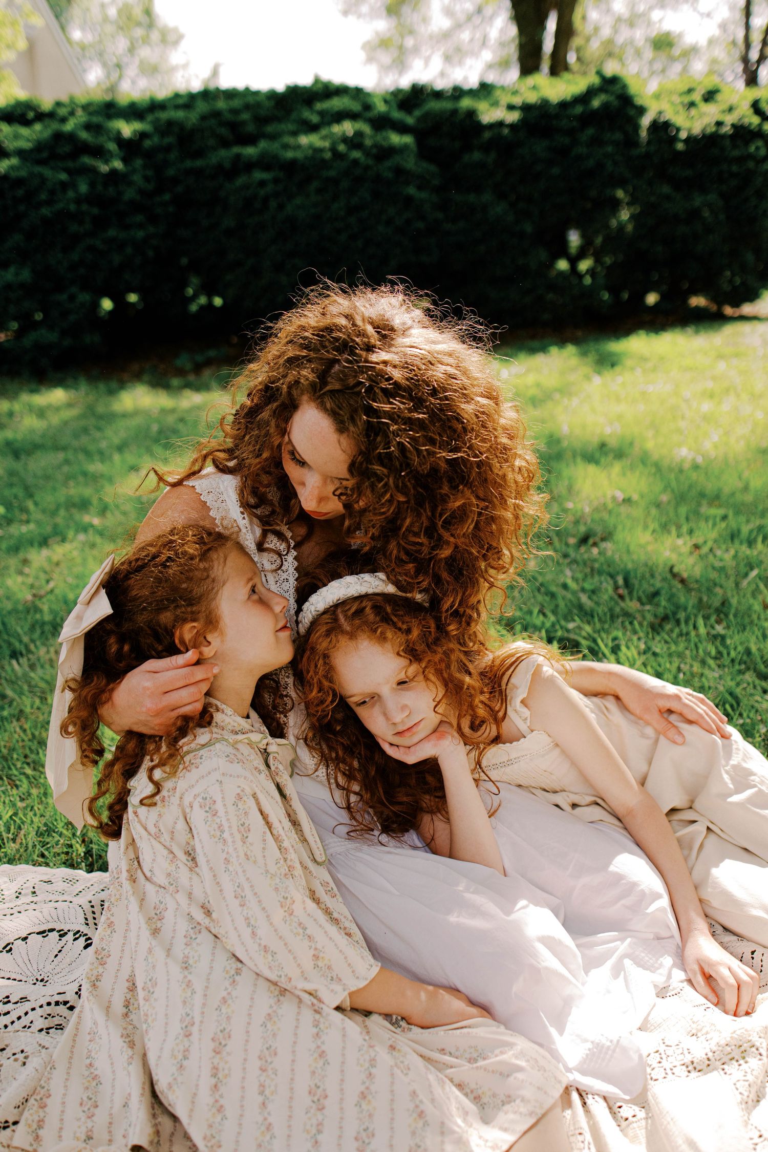 Curly-haired mother in vintage-style dresses cuddles two young daughters on a lace blanket in a sunny green park.