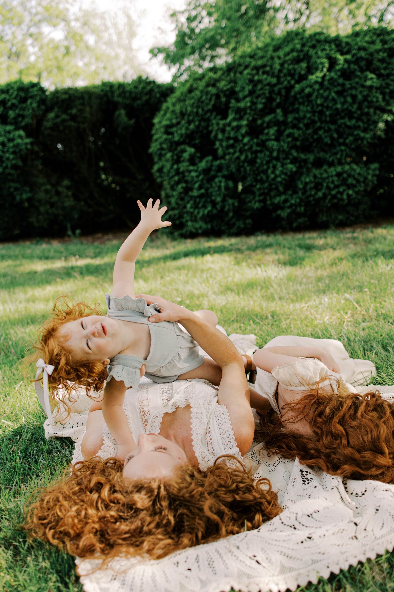 Red-haired mother and curly-haired toddler laughing together on a lace blanket in a sunny green park.