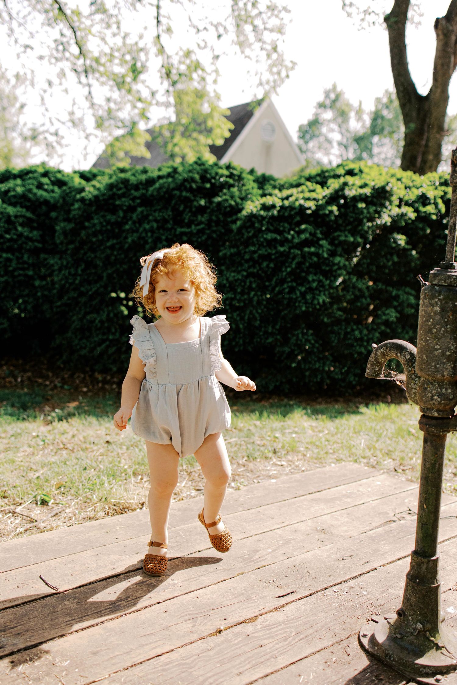Smiling toddler with curly red hair and a white bow runs across a wooden deck in a gray romper near an antique water pump.