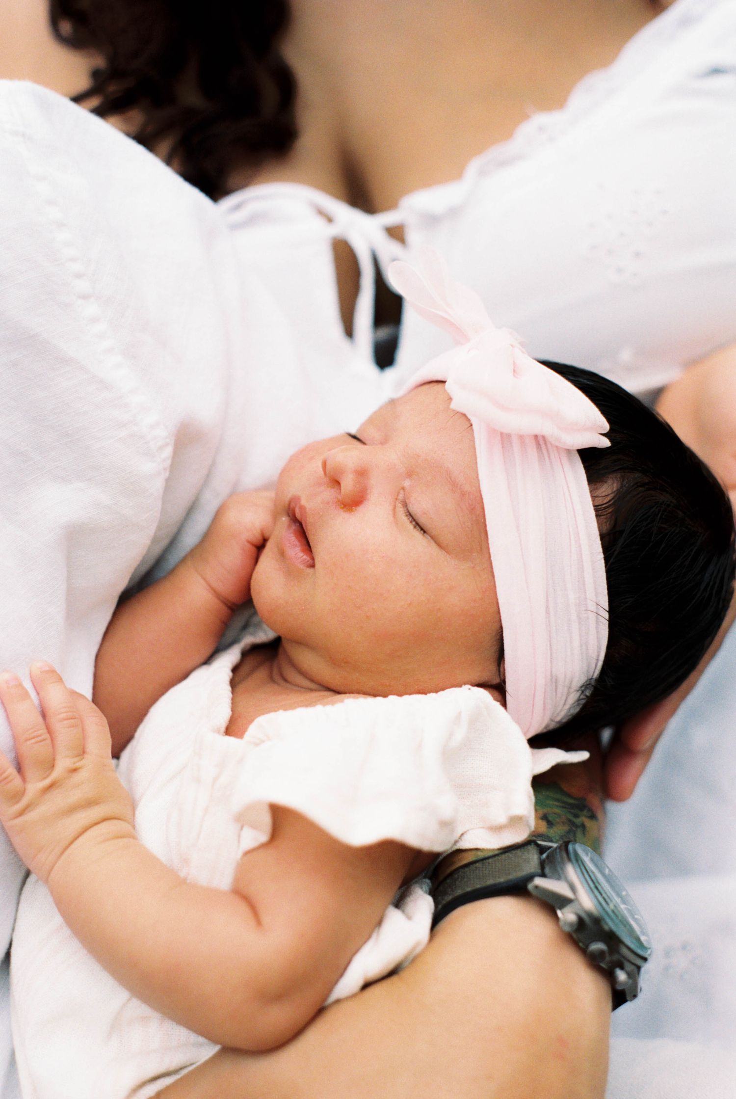 Sleeping newborn baby girl in white outfit and pink bow headband cradled in mothers arms.