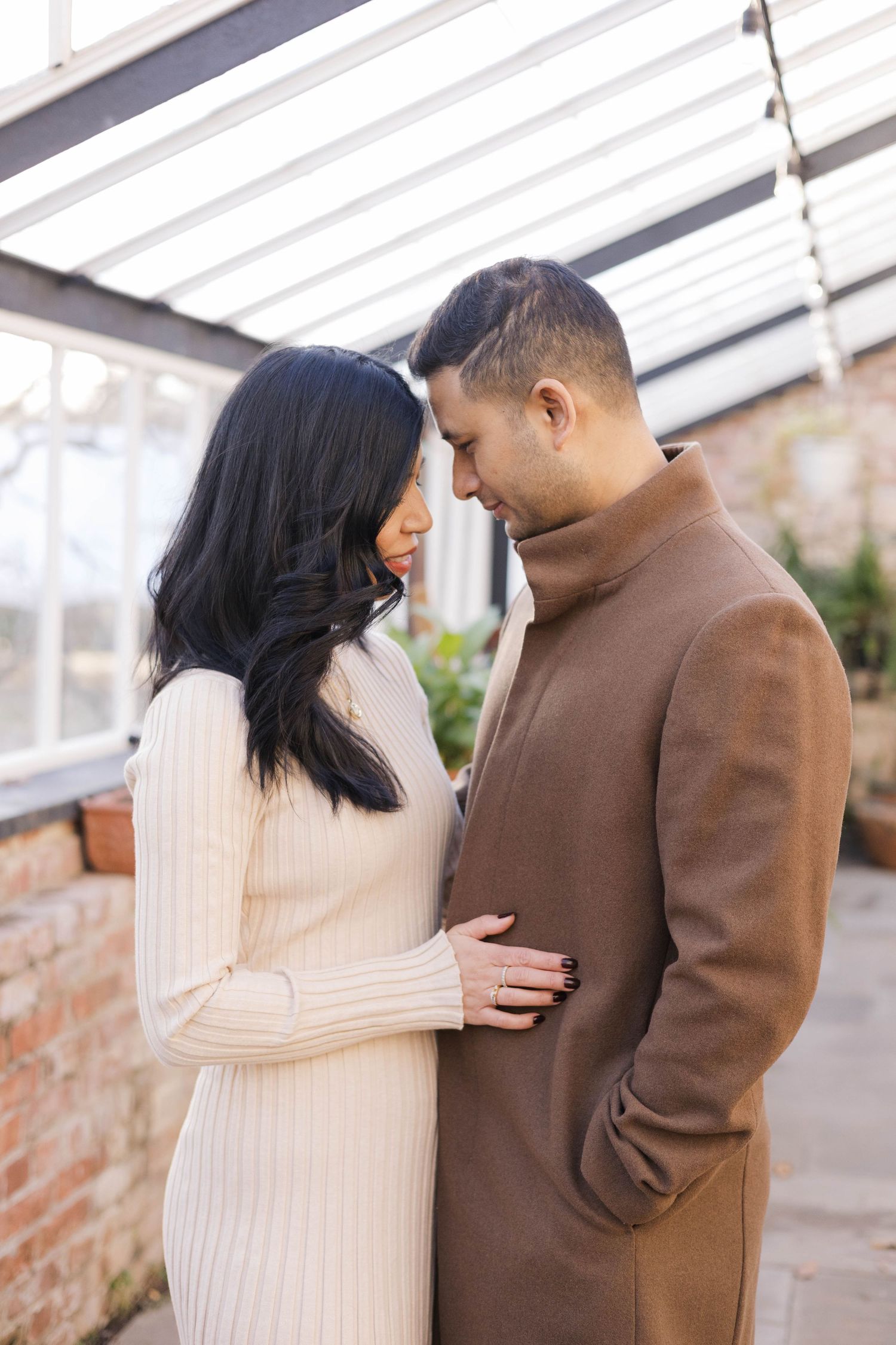 Couple in winter coats sharing intimate moments against a white greenhouse backdrop.
