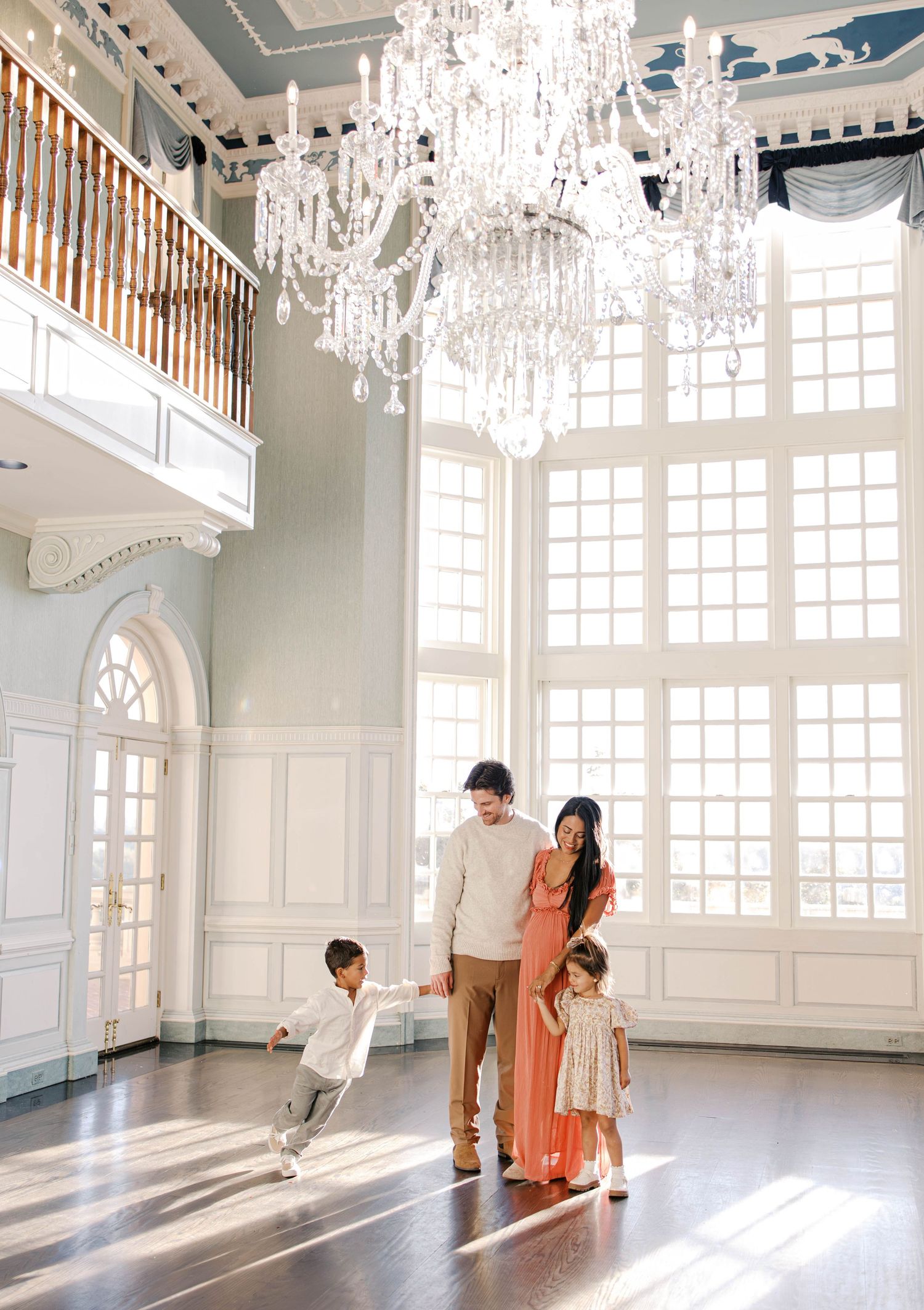 Family of four in coordinated neutral outfits stands in a grand sunlit ballroom with a crystal chandelier while a young boy dances nearby.