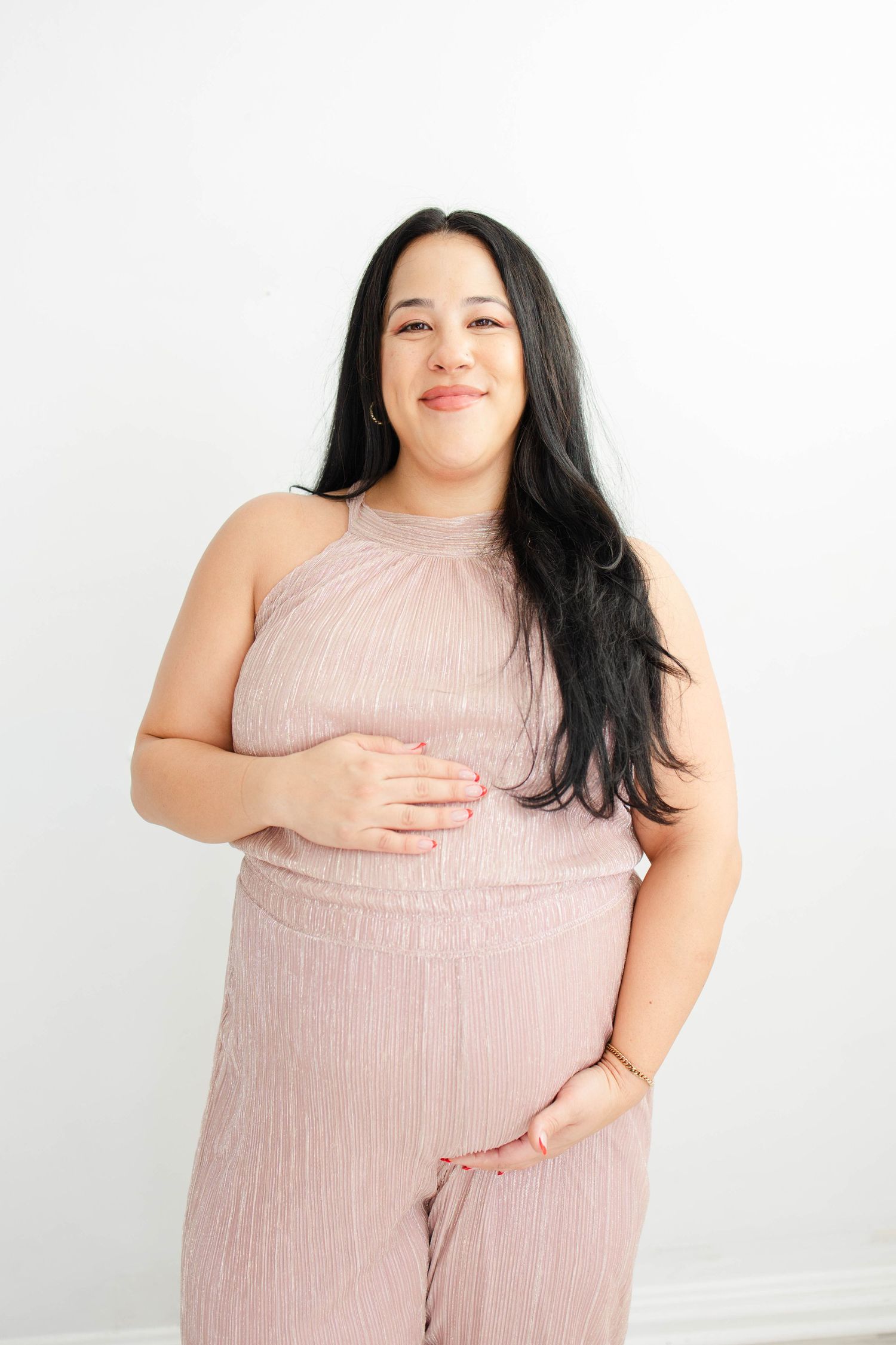 Woman in blush pink halter dress poses with hands on belly against white wall.