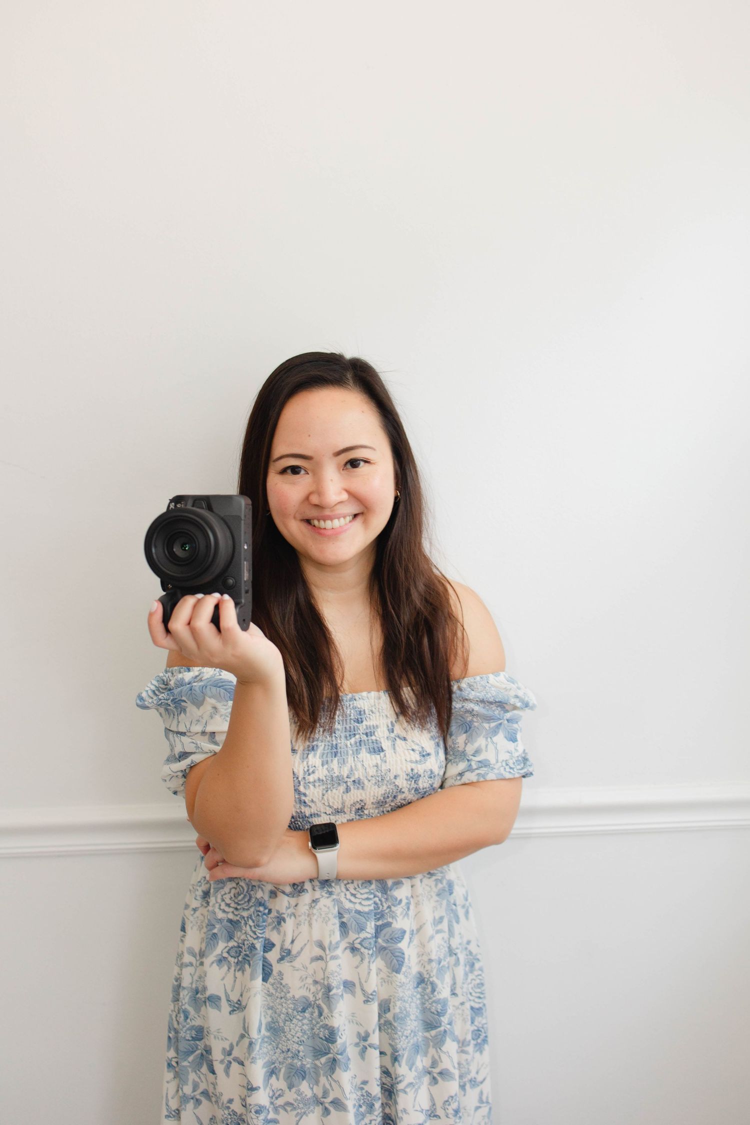 Photographer in floral dress poses with camera against white wall.
