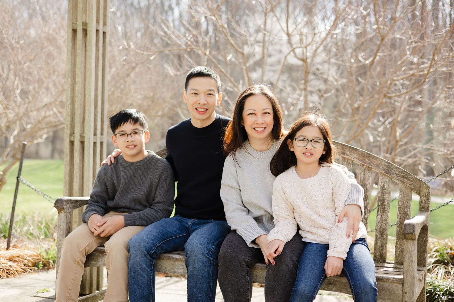 Family of four smiling together on a wooden bench outdoors in a park during winter.