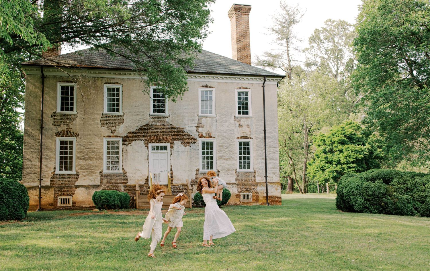 Three girls in white dresses dance on a sunlit lawn in front of a historic brick manor house surrounded by tall trees.