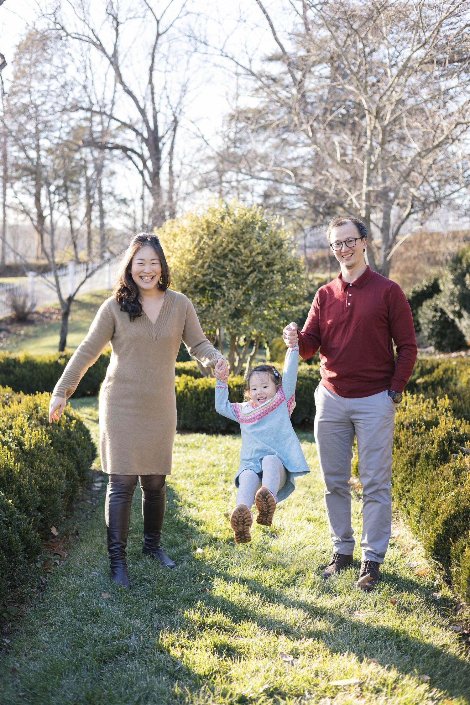Family members playing and jumping along a garden path surrounded by evergreen bushes.