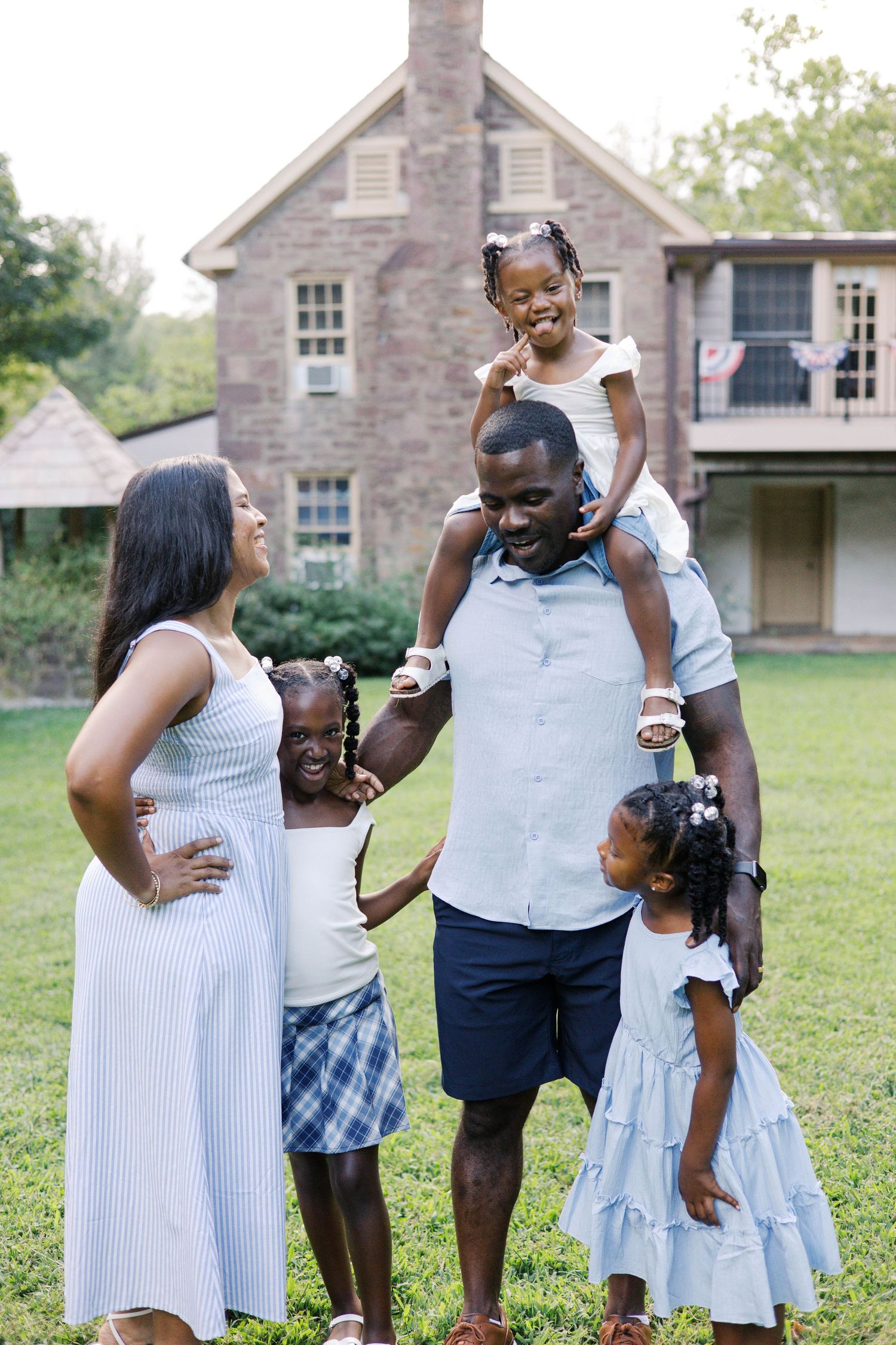 A joyful Black family poses outdoors with three young daughters, dad carrying one on shoulders, in front of a brick home.