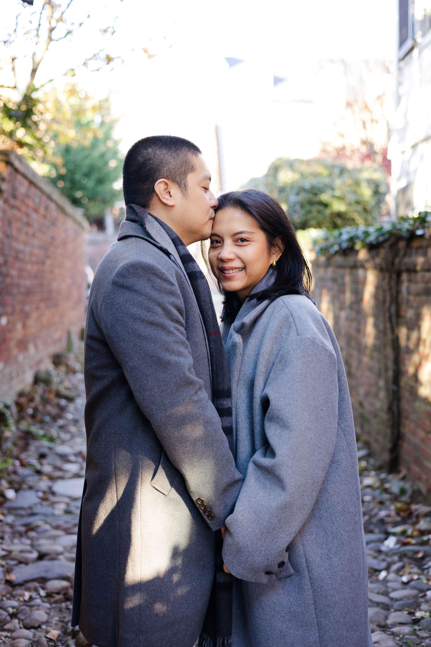Couple in winter coats sharing playful moments and laughter against a rustic brick wall backdrop.