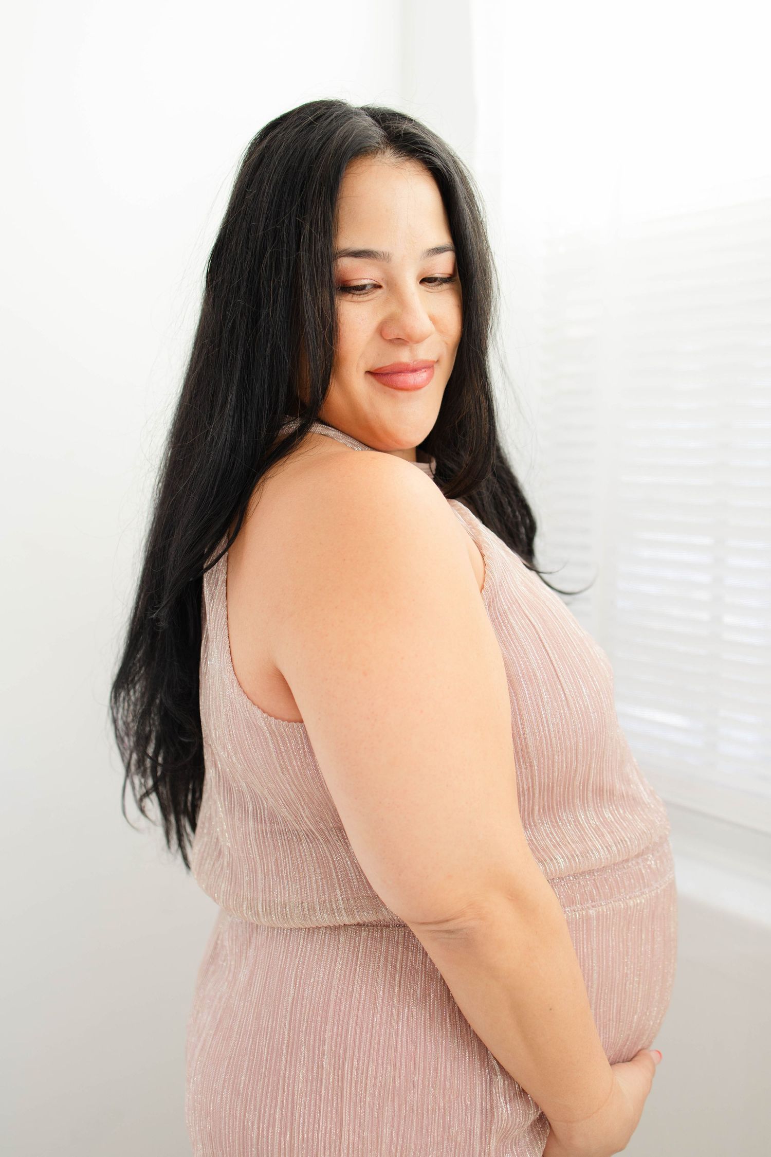 Side view of woman in pink dress showing baby bump against bright window.
