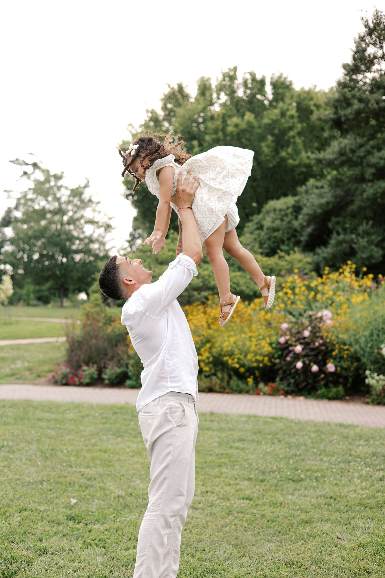 Father in white outfit joyfully tosses young daughter in white dress high in the air in a lush green park.