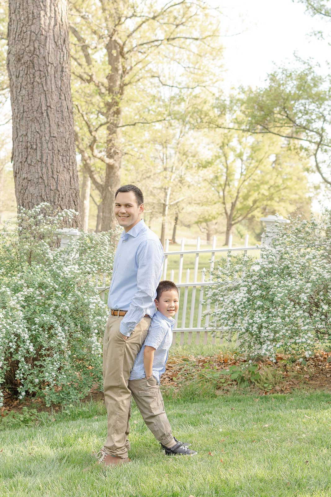 A father and young son pose together smiling in a lush green garden with white flowering bushes and trees.