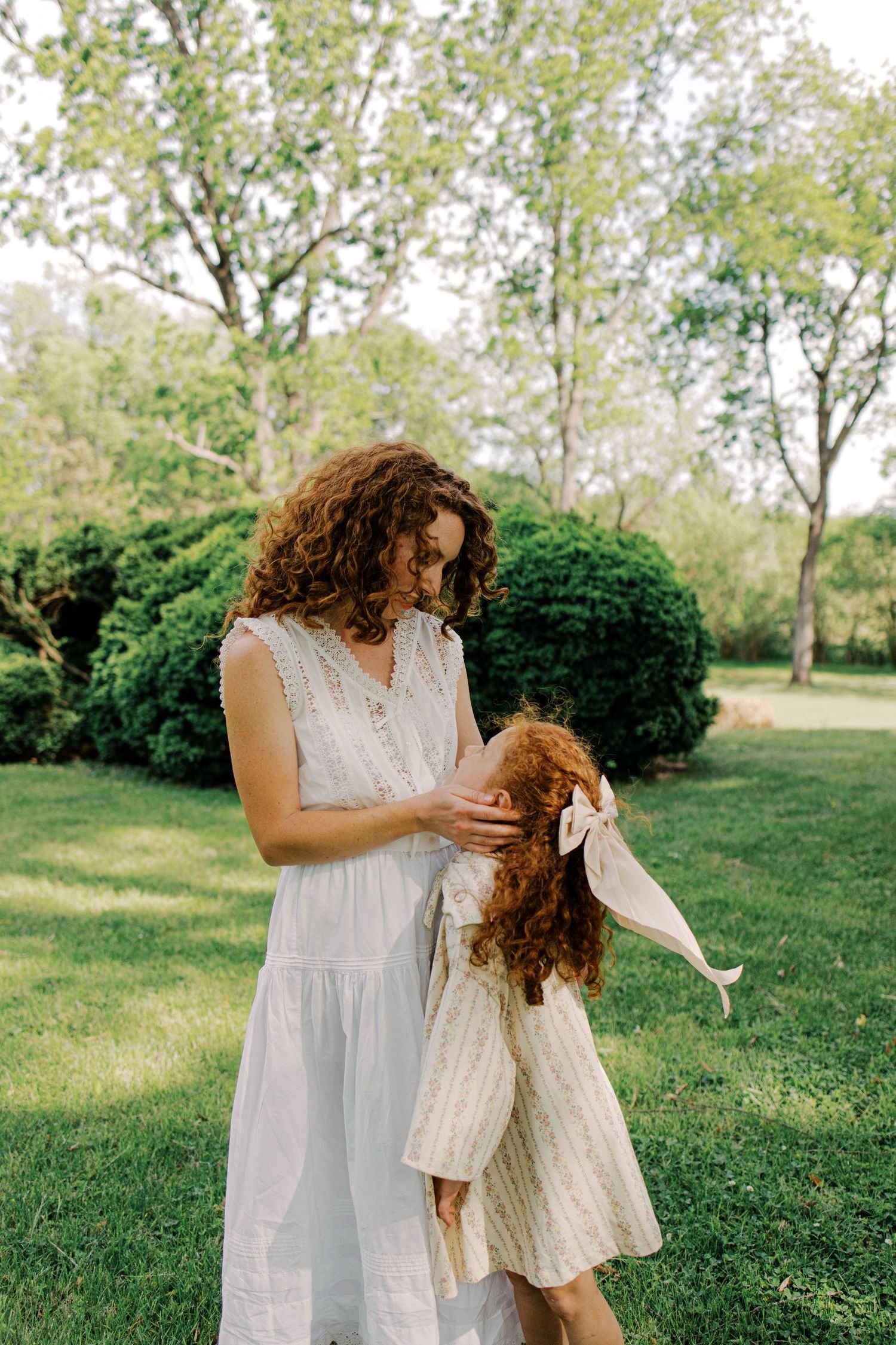 A curly-haired woman in a white sundress gently touches a little girl in a striped dress and bow in a sunny green garden.