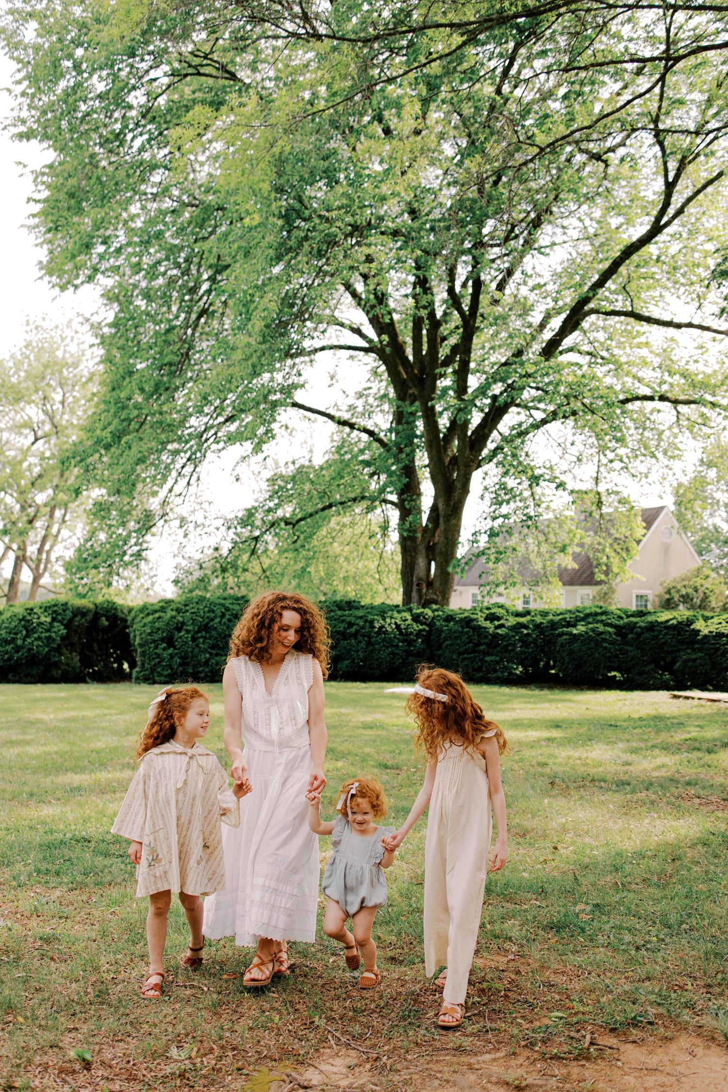 A woman and three young girls in light dresses hold hands while walking across a sunny grassy lawn beneath a large tree.