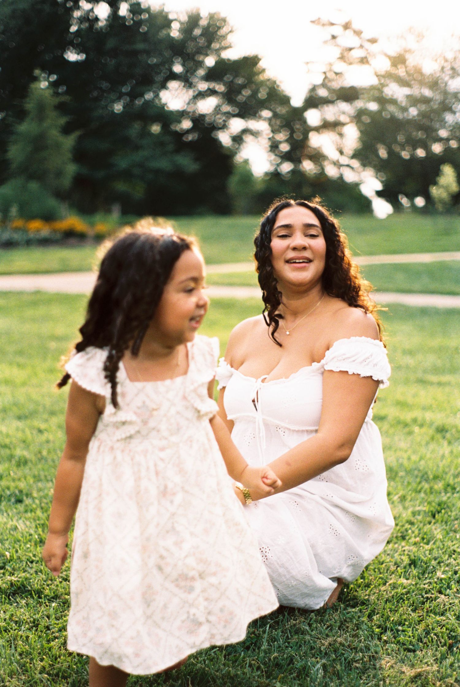 A woman in a white dress crouches smiling at a young girl in a white dress on green grass in a sunny park.