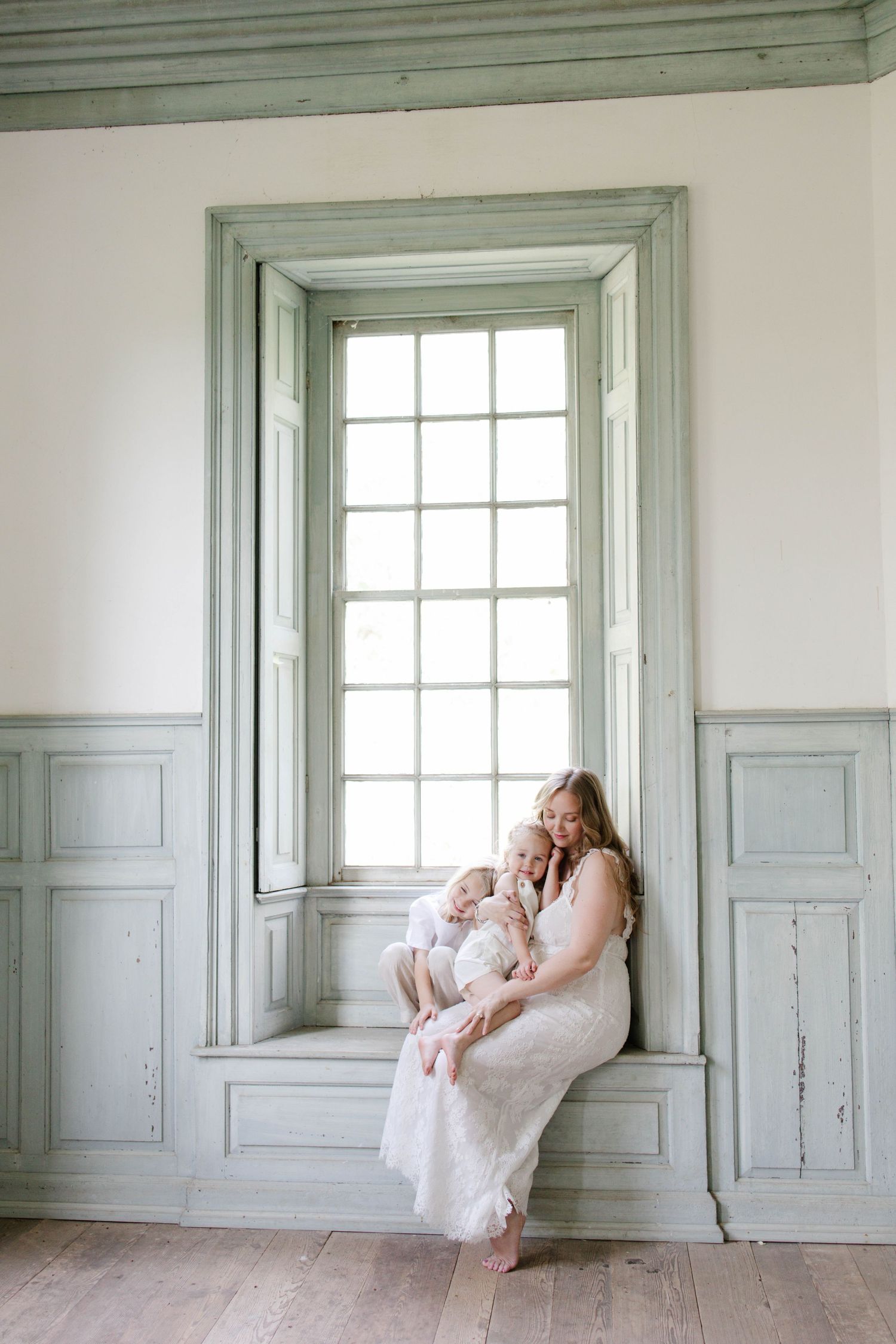 A mother in a white dress sits on a window seat holding two young children in a elegant room with sage green paneling.