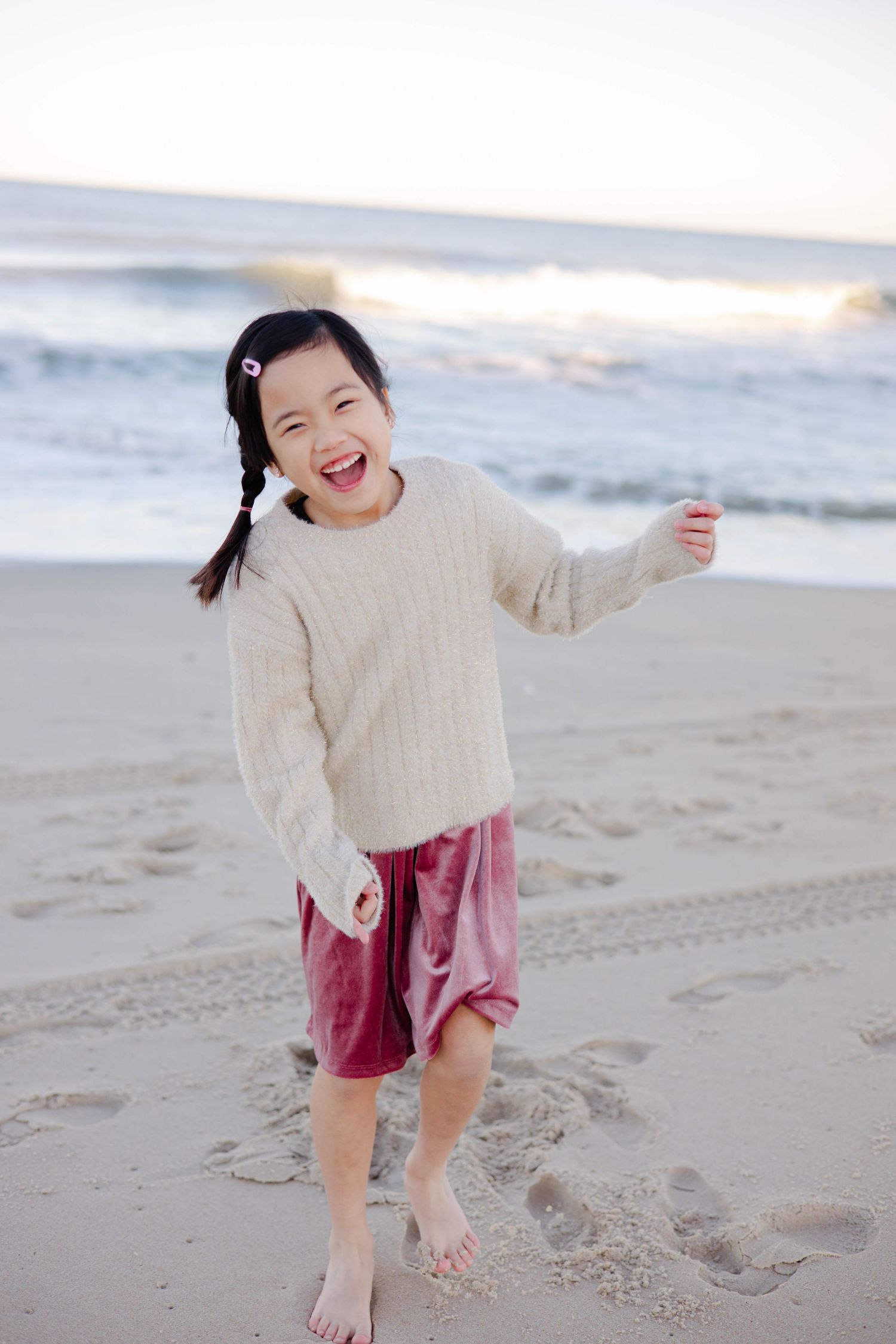 A joyful child in a cream sweater plays barefoot on the beach.