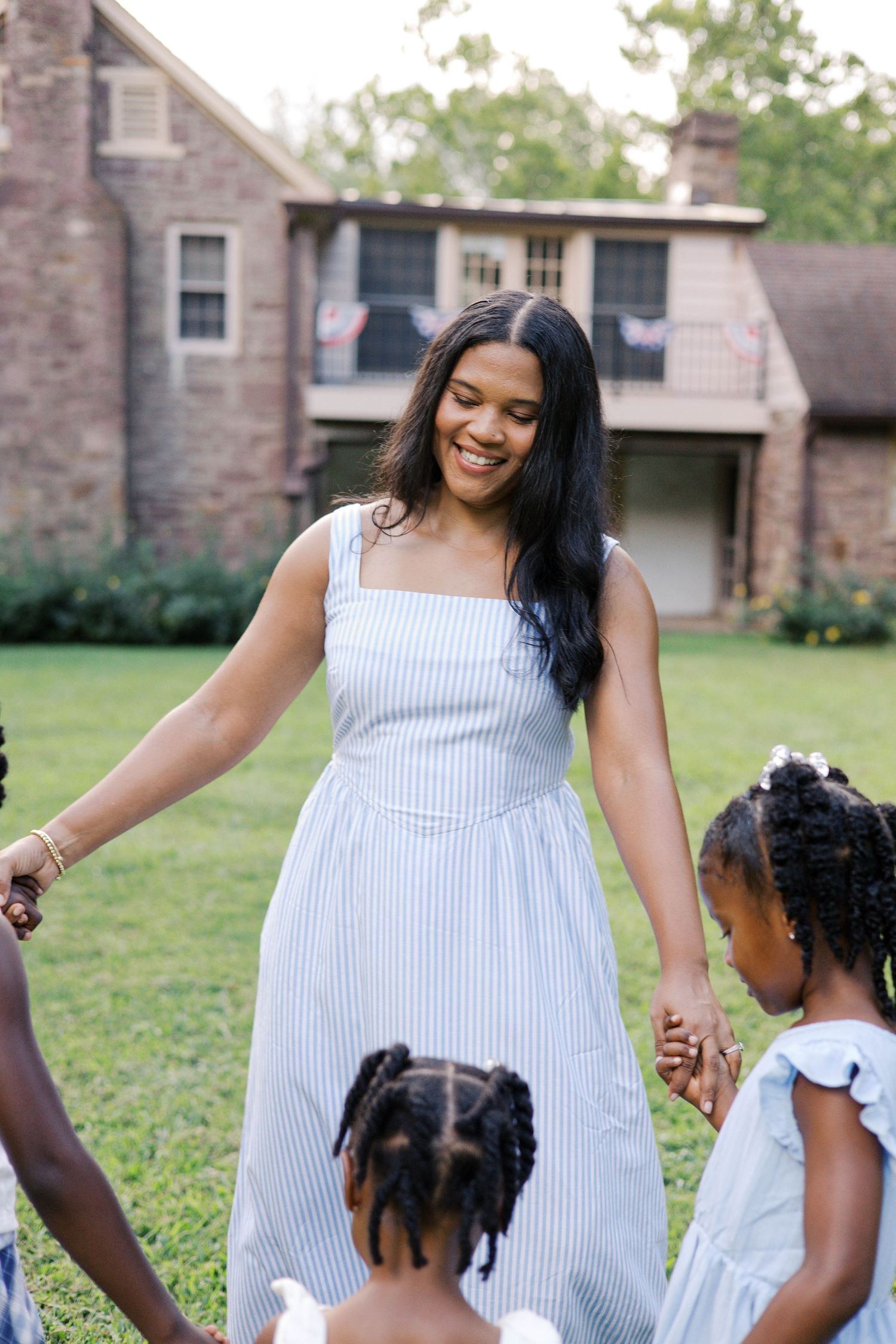 A smiling woman in a blue striped dress holds hands with children on a green lawn outside a brick house.