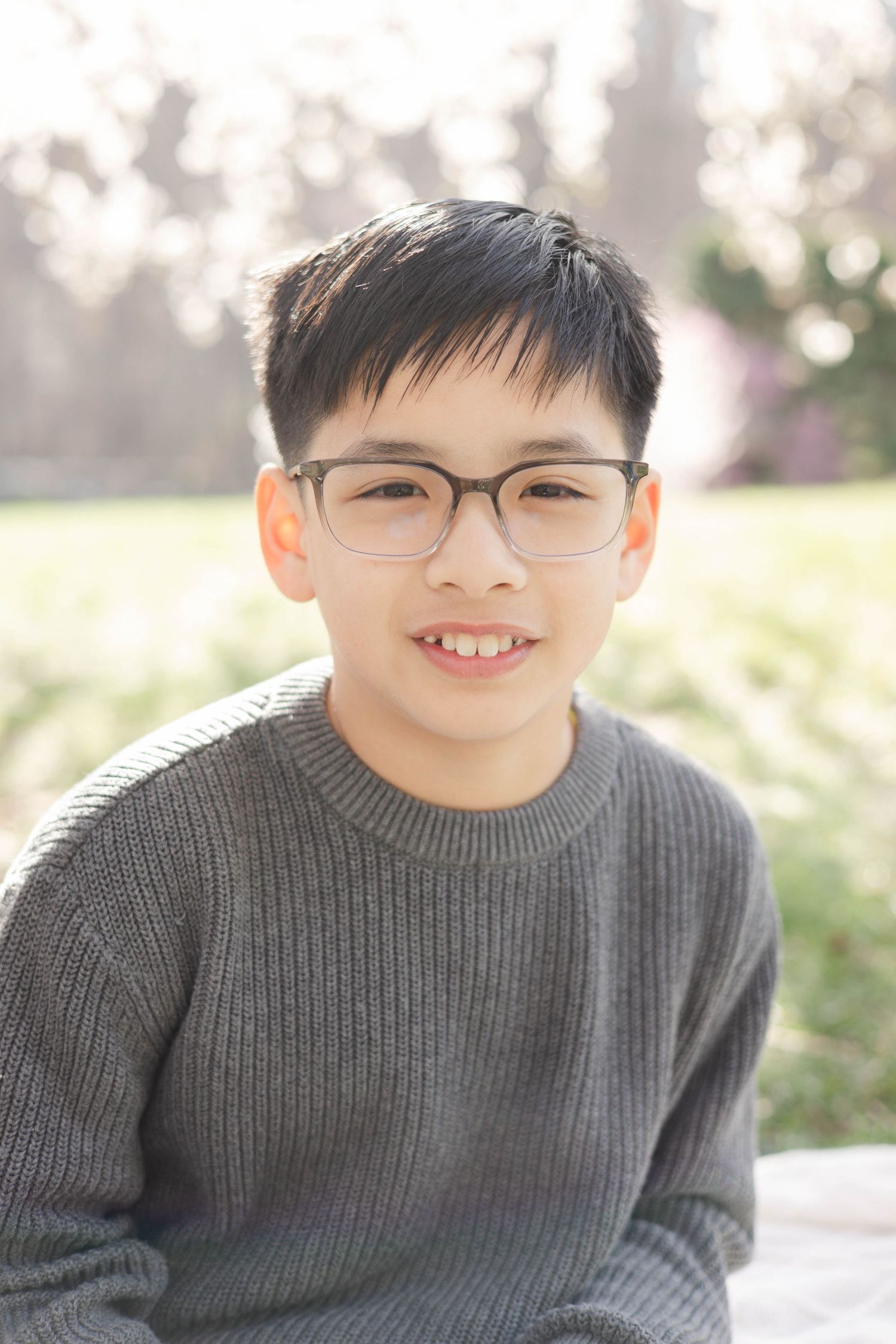 Smiling young boy with glasses wearing a gray sweater, posing outdoors in a sunny park setting.