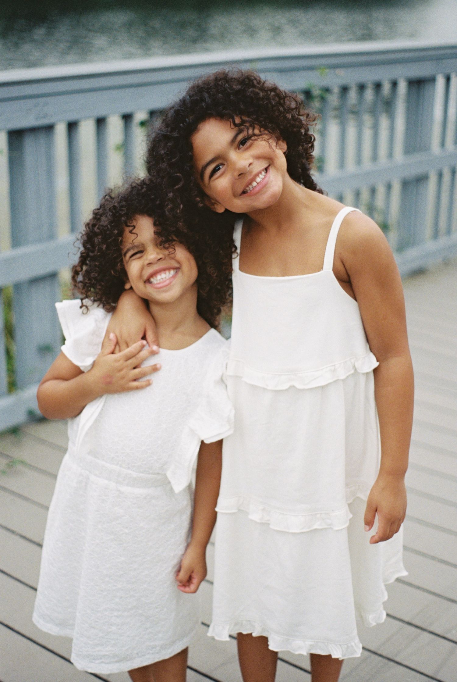 Two smiling girls in matching white dresses pose together on a wooden deck, both with curly dark hair.