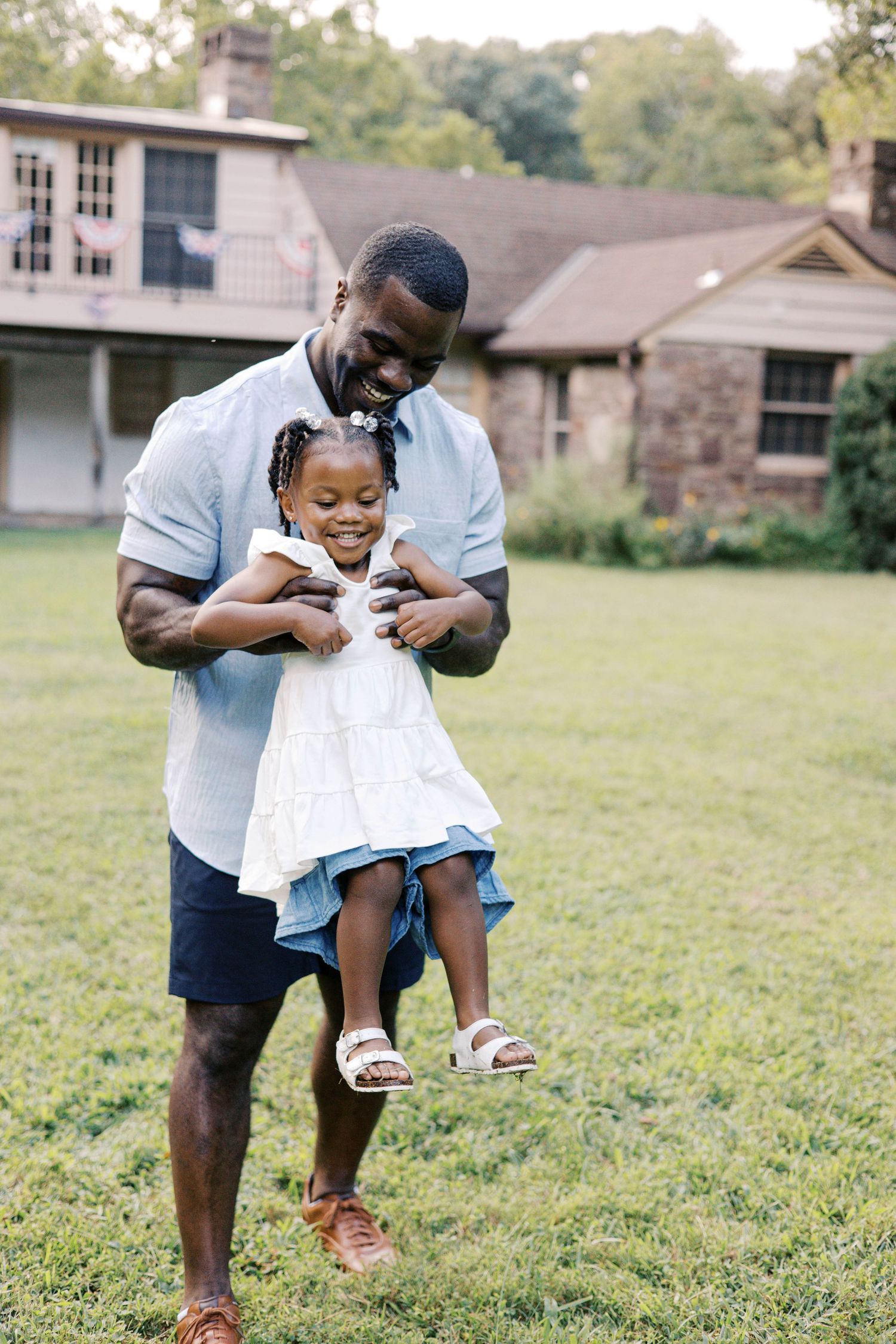 A joyful father lifts his smiling young daughter in a white dress outdoors on green grass near a stone house.