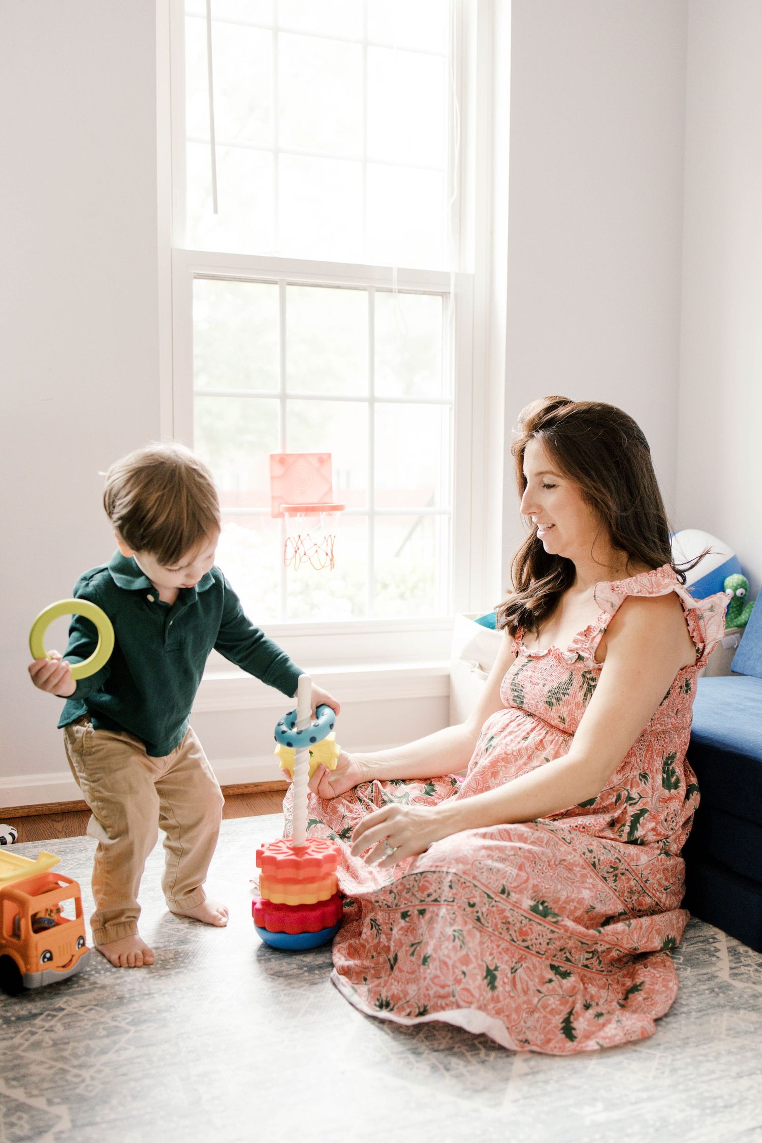 Pregnant woman in floral dress plays with toddler boy stacking rings on nursery floor by bright window.