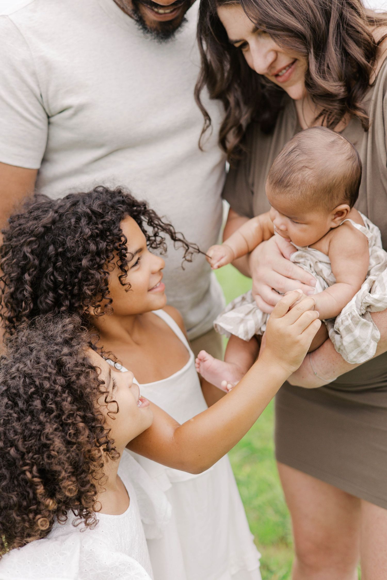 A joyful family gathers outdoors, with parents and two curly-haired girls admiring a newborn baby.
