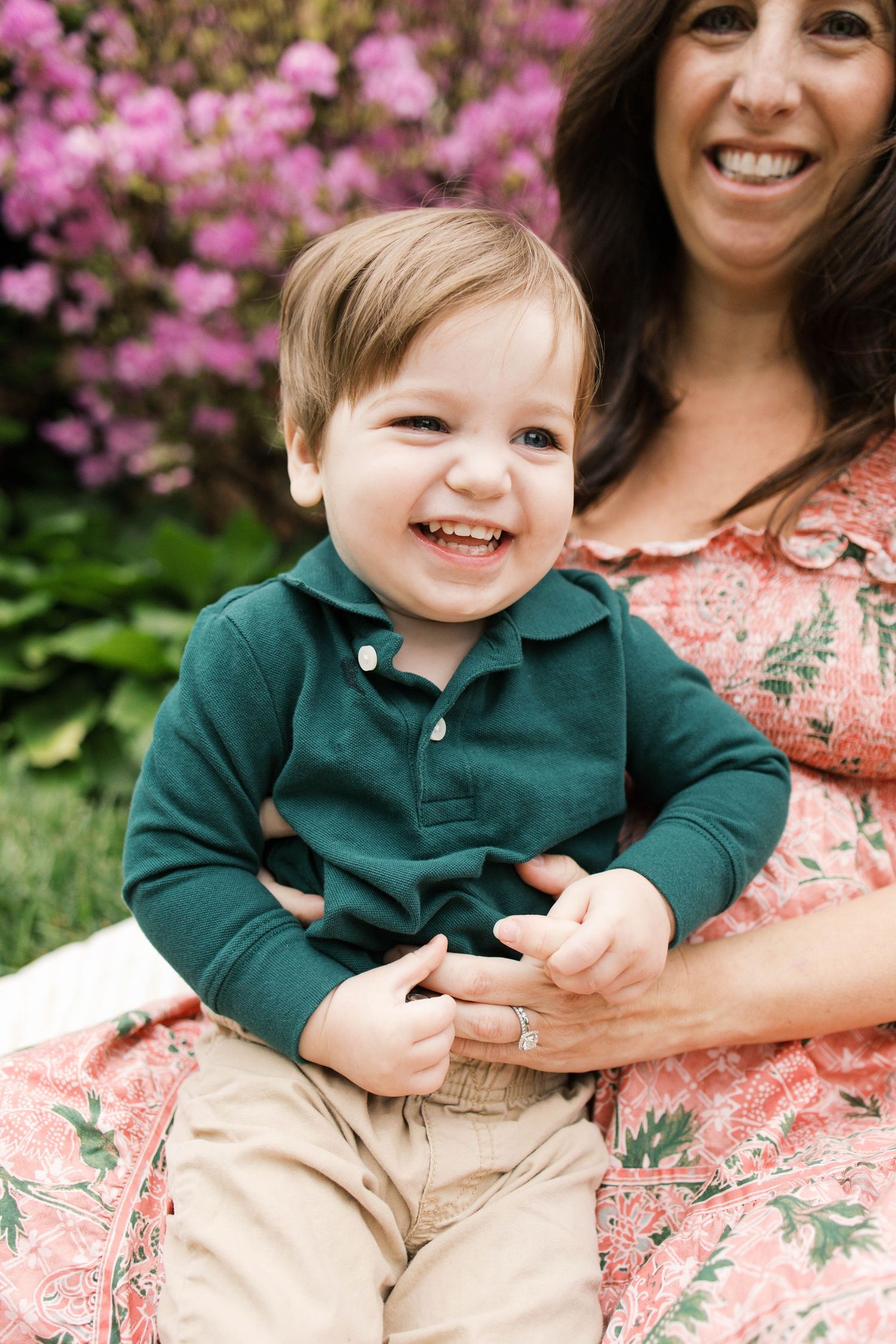 Smiling toddler boy in green polo shirt sitting on mother's lap outdoors with pink flowering bushes.