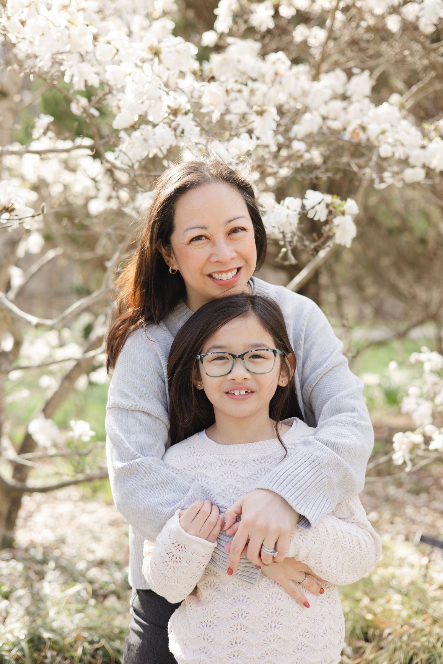 A woman and young girl with glasses smile together in front of white blooming magnolia trees in spring.