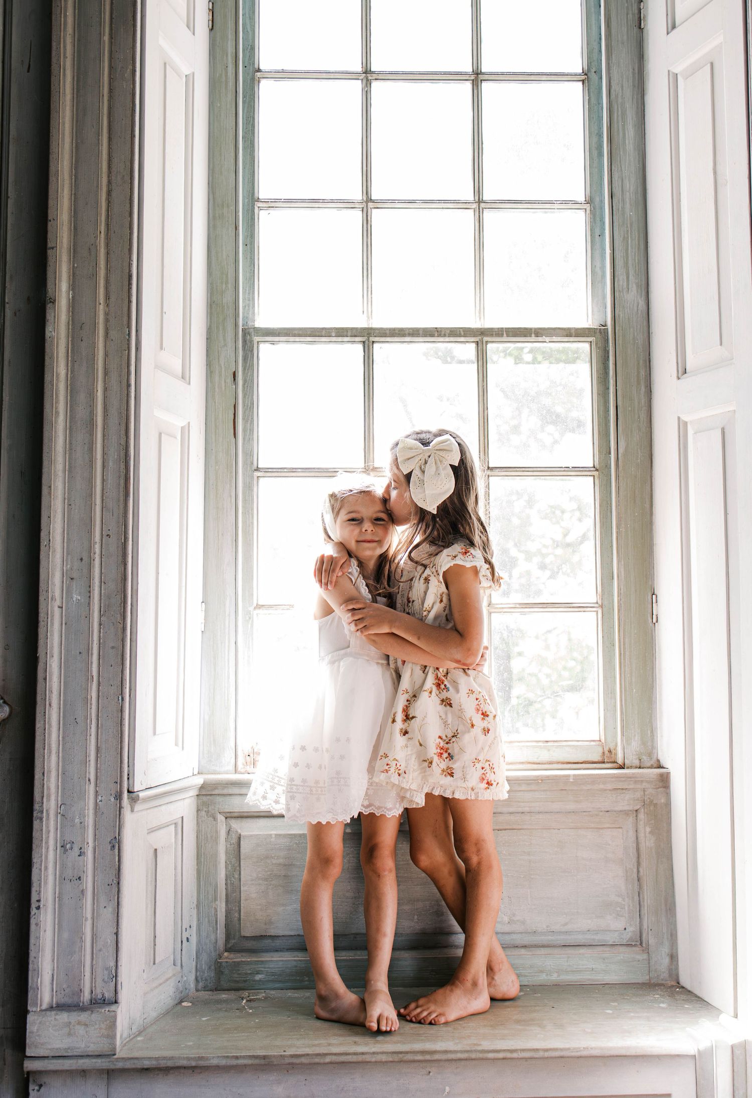 Two young girls in floral and white lace dresses embrace barefoot on a rustic window sill as one kisses the other's cheek.