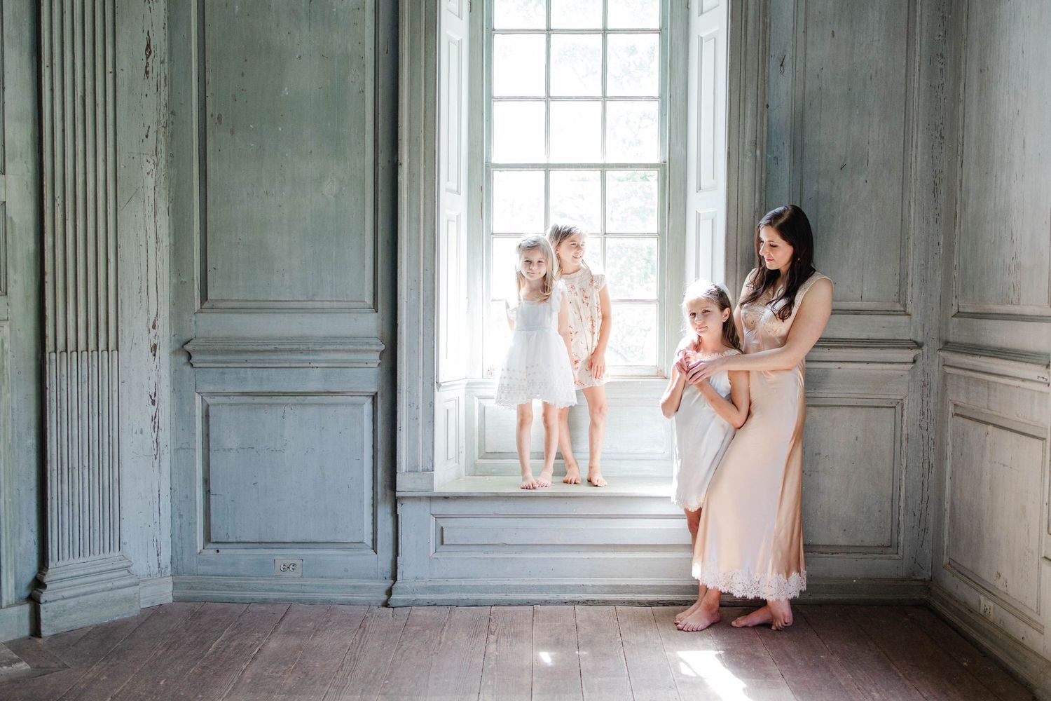 A mother holds her baby while two young girls play by a large window in an elegant, softly lit room.