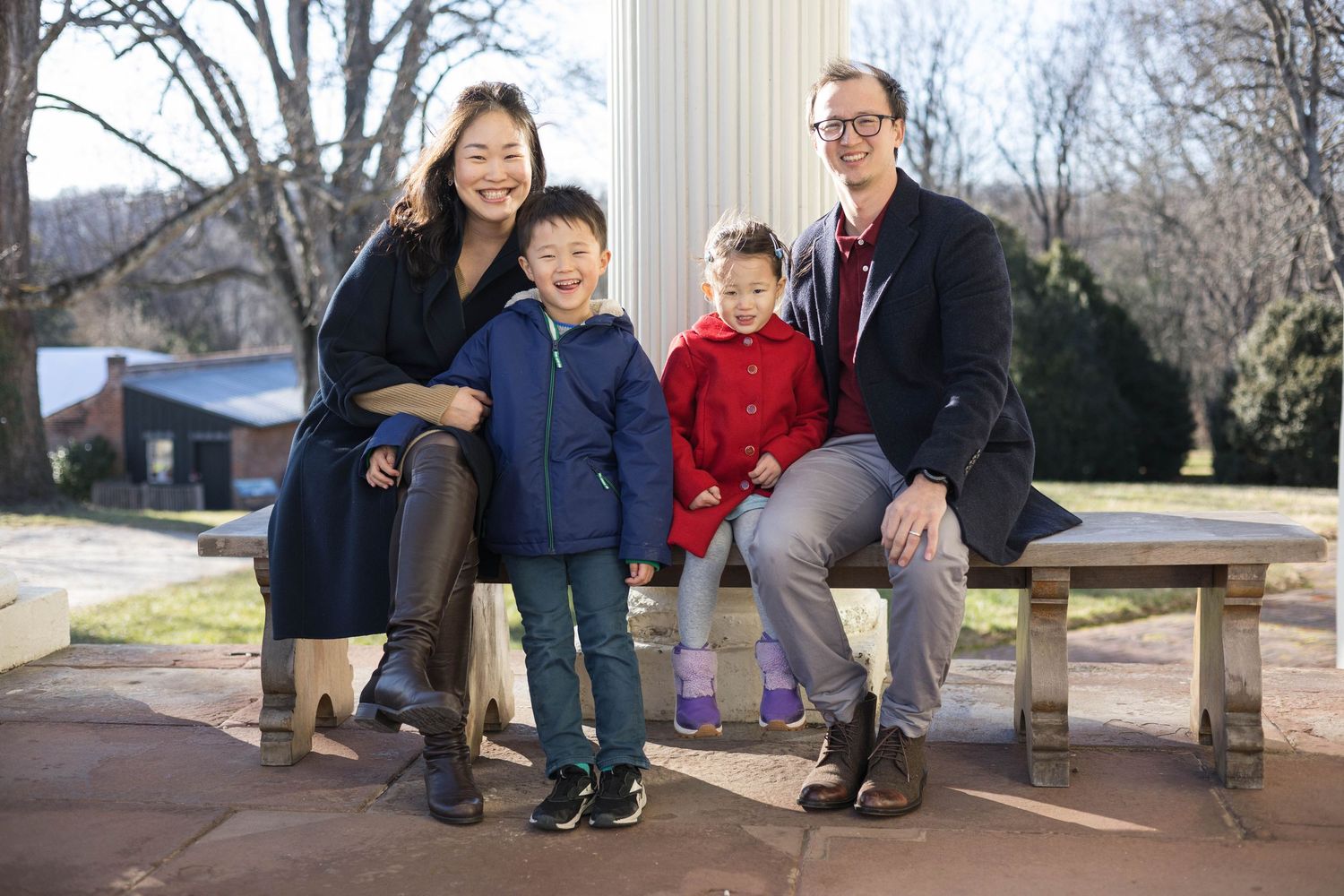 Family sitting together on wooden bench by white column outdoors.