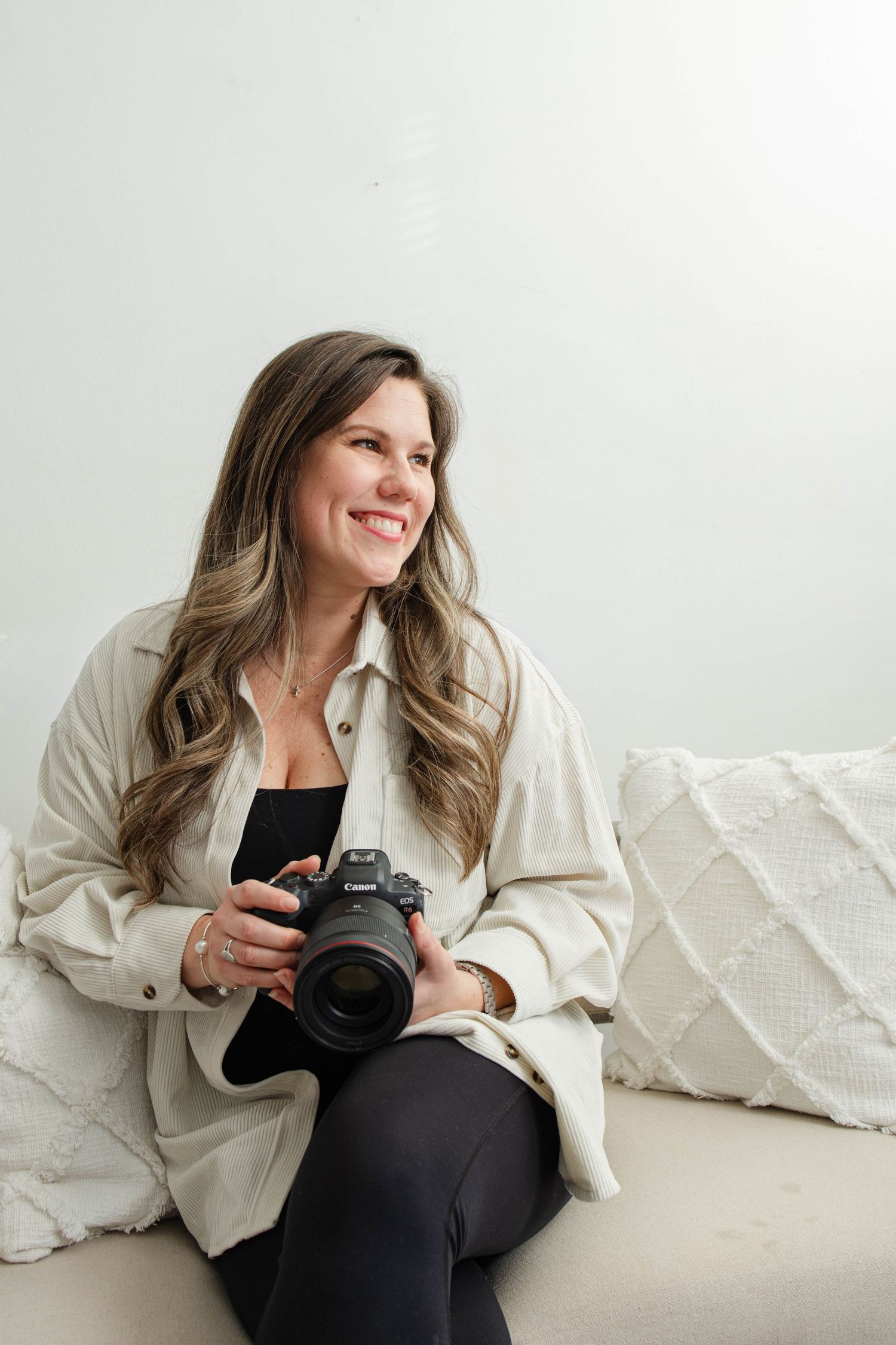 Photographer in cream jacket poses with camera on white textured couch.