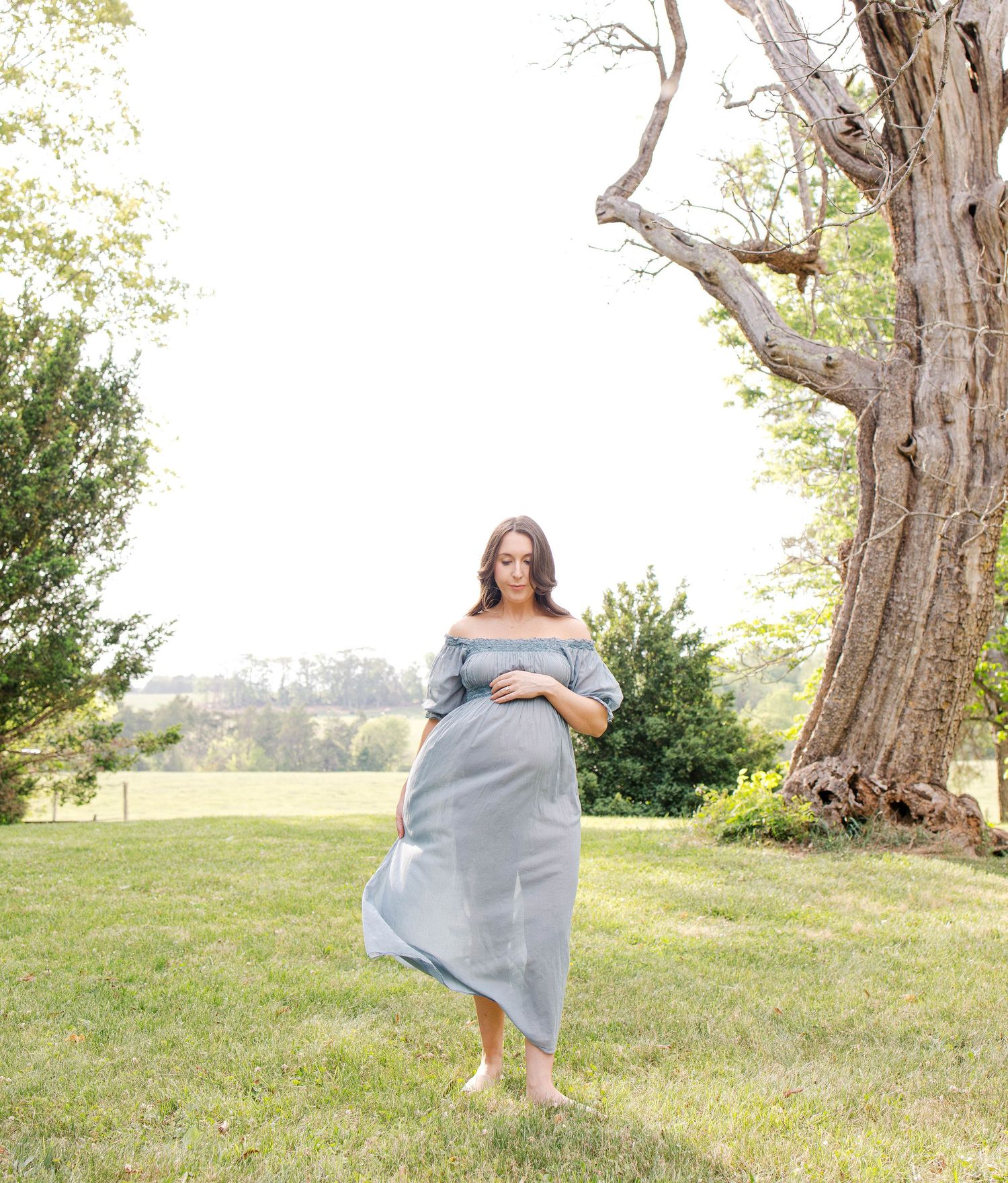 Pregnant woman in flowing blue off-shoulder dress standing barefoot on green grass near large tree during outdoor maternity photoshoot.