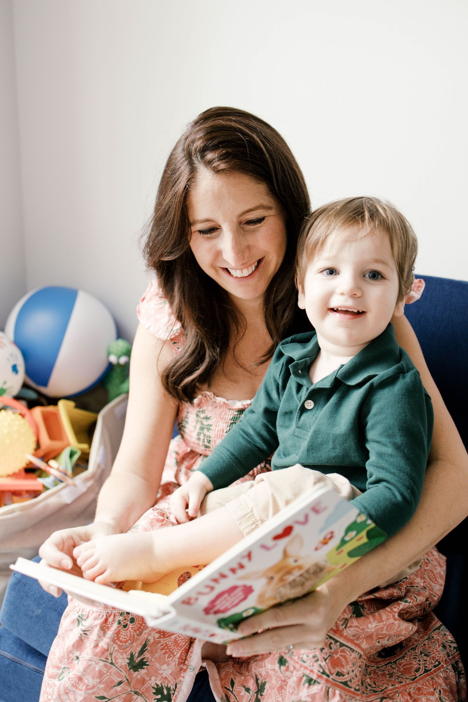 Smiling mother reading a colorful picture book with her happy toddler boy on her lap in a playroom.